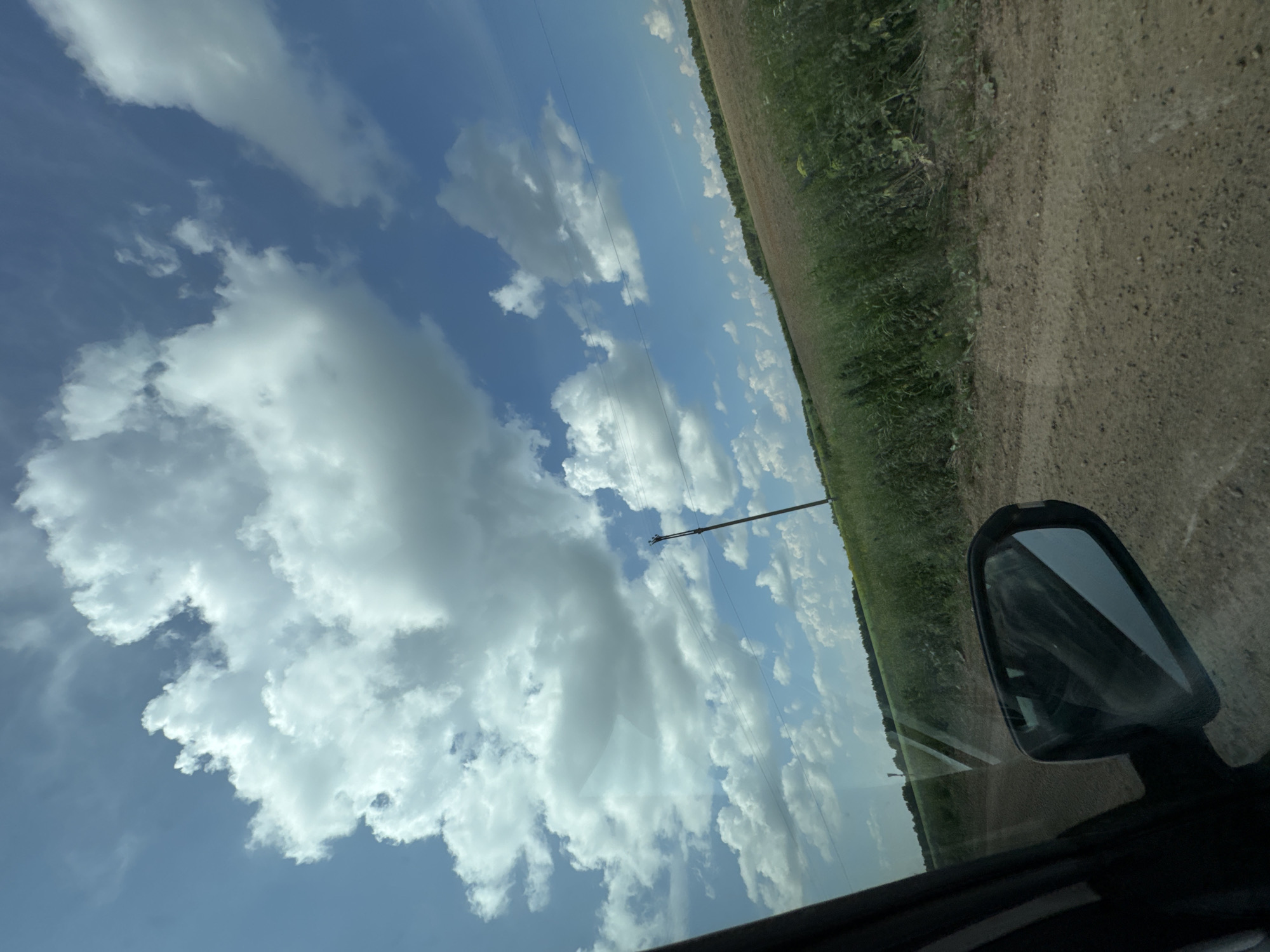 A side view of a country road in the city of Vassyata, Russia, taken from inside a vehicle. The sky is bright blue with fluffy white clouds. A dirt road is visible in the foreground, with a field of green grass and crops to the right. A single telephone pole stands in the distance. The side mirror of the car is visible in the lower right corner of the image. The overall scene depicts a rural landscape on a sunny day.