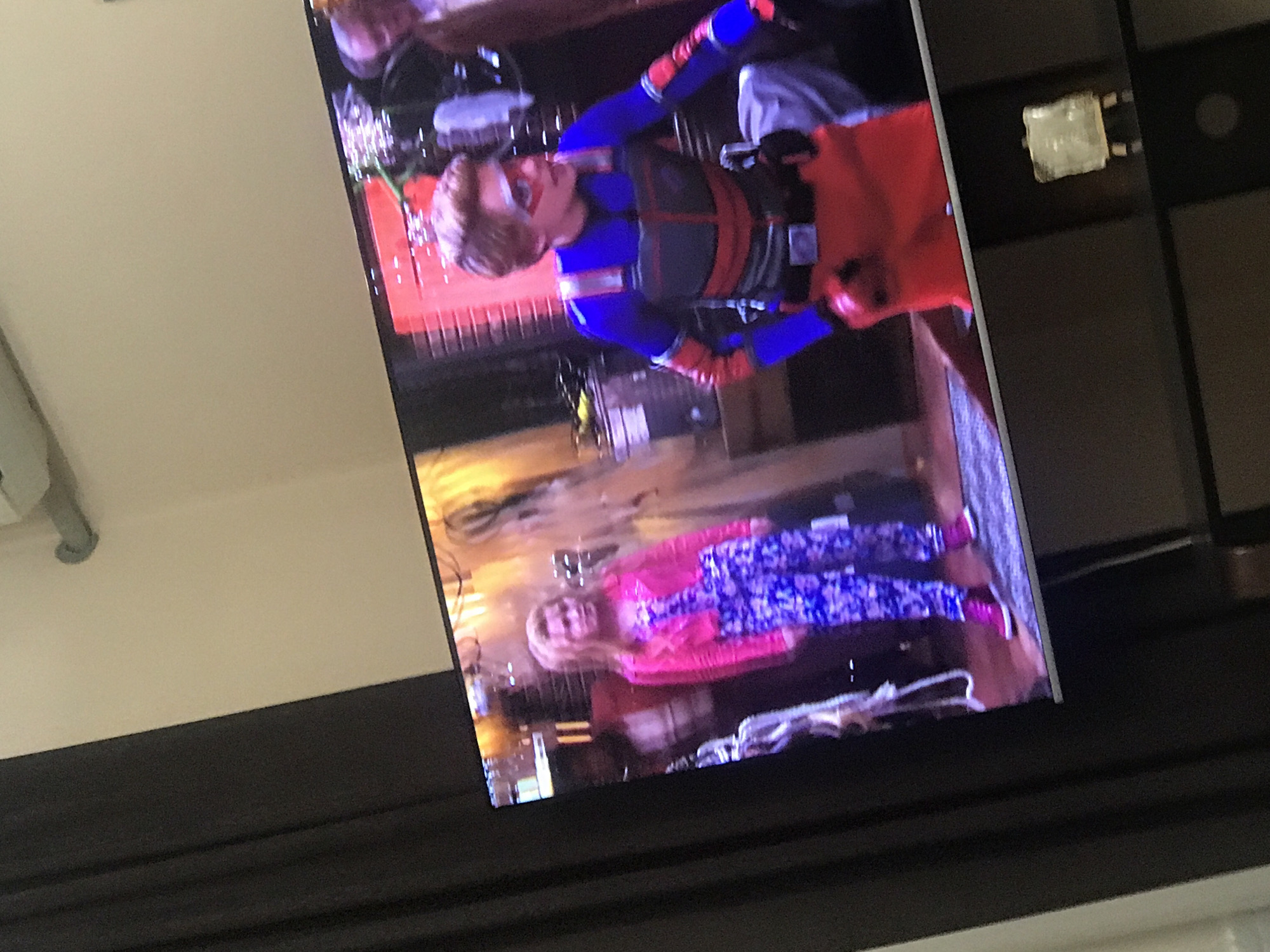 The image shows a television screen displaying a scene with two characters. One character is a blonde-haired male wearing a superhero-style costume with blue and red accents, including a mask and orange gloves. The second character is a blonde-haired female wearing a pink top and blue patterned pants. The scene appears to be indoors, possibly a living room or entertainment space. The television is placed in front of a dark-colored wall.
This image does not depict a location in Yerevan, Armenia.