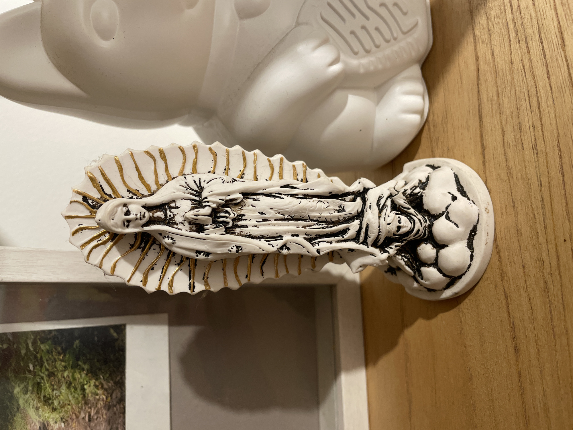 The image shows a close-up view of a white and gold Our Lady of Guadalupe statue resting on a light-colored wooden surface. A partial view of a white figurine is visible at the top of the image. Part of a white picture frame with a visible image is also at the bottom of the image. The lighting appears to be from an overhead source. The location is not discernible from the provided image.