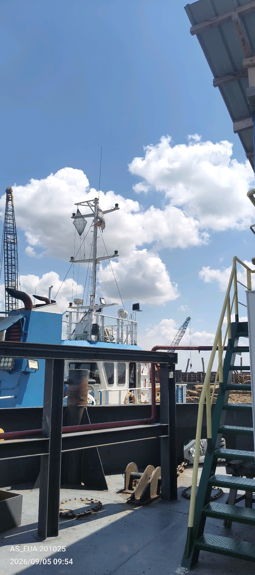 A daytime, outdoor, medium shot captures a blue and white boat docked near a facility with a bright blue sky dotted with fluffy white clouds. In the foreground, a dark metal railing is visible, with green and yellow metal stairs ascending to the right. The deck area is concrete, showing some wear and tear, with a metal hatch cover and two large metal anchors.
Further back, the boat's superstructure, including a white cabin with windows and a mast with antennas and a diamond-shaped black object, is prominent. A large industrial crane with a lattice structure stands to the left of the boat, partially obscured. To the right of the boat and behind it, another crane is visible, along with stacks of logs and other industrial equipment. The scene appears to be a shipyard or port facility. The timestamp on the image indicates it was taken on September 5, 2026, at 09:54.