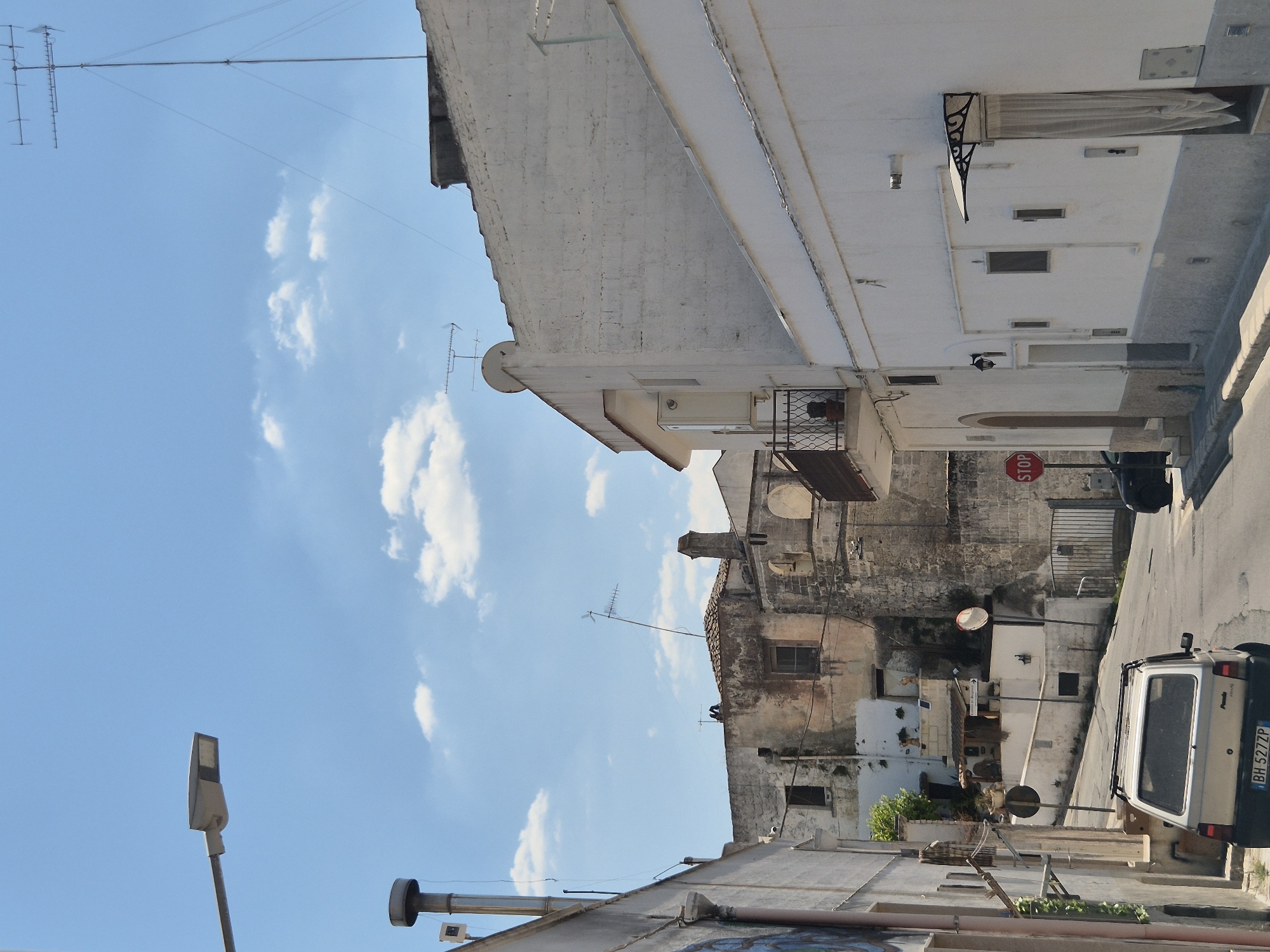 The image captures a street scene in Laterza, Italy, on a clear, sunny day with scattered clouds. A narrow, paved street winds uphill, bordered by buildings of varying architectural styles. On the left, a light-colored building features a street lamp and what appears to be an entranceway with graffiti. Further up the street, older stone buildings exhibit signs of age and wear, with visible cracks and weathered facades.

In the foreground, a silver Fiat Panda is parked on the right side of the street, facing uphill. Its license plate reads "BH 527ZP". To the right of the Panda, a black car is partially visible.

Dominating the right side of the image is a white, two-story building with a white awning over a doorway. The wall of this building is made of rough-hewn white blocks. Several windows are visible on this building, some with protective grates or coverings.

Antennas are prominent on the rooftops of several buildings, indicating modern infrastructure within the older urban fabric. A red "STOP" sign is visible on the right side of the street, near the black car. The overall impression is of a well-established, possibly historic, Italian town with elements of both old and new architecture.