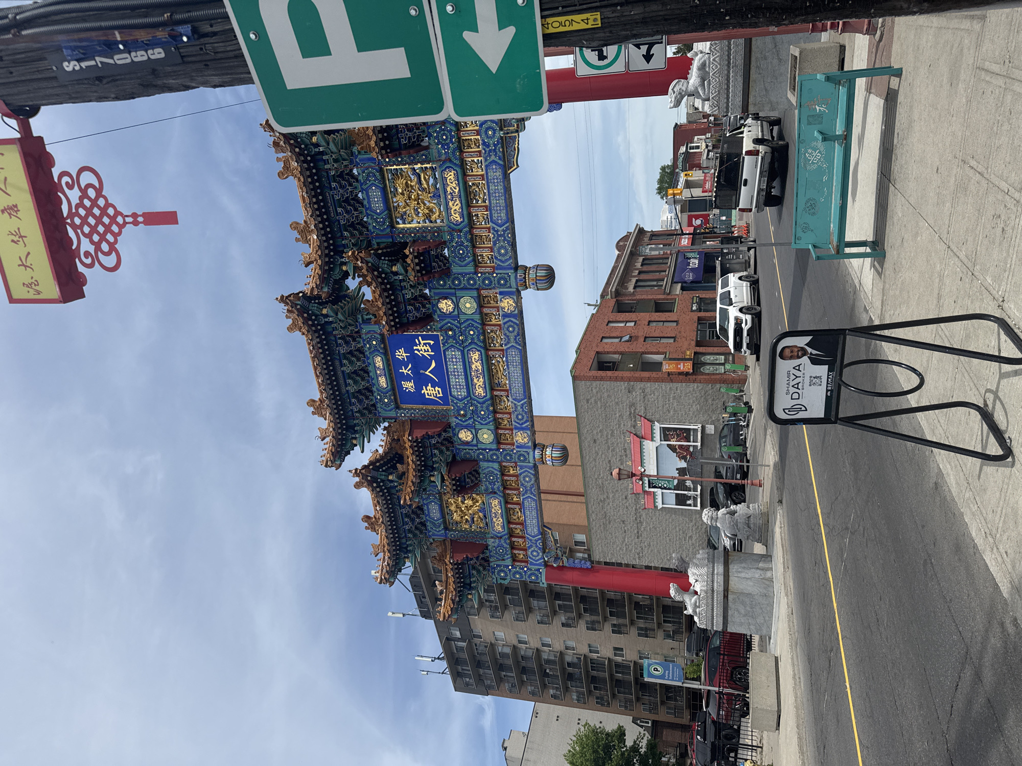 The image displays a vibrant urban street scene, rotated 90 degrees clockwise. The central focus is an ornate, multi-colored Chinese gate (Paifang) with intricate blue, gold, and red decorations. A central plaque on the gate is inscribed with Chinese characters "渥太華 華人街," identifying the location as "Ottawa Chinatown." Banners with "恭賀新禧" (Happy New Year/Season's Greetings) are also visible. A red traditional Chinese knot decoration hangs near the gate.
The street, appearing vertical due to the rotation, is paved with a yellow dividing line. Buildings of varied architecture, including brick and concrete structures with balconies, flank the street. Several vehicles, such as a white pickup truck and a white SUV, are present on the road.
On the right side, a light blue-green public bench is situated on the sidewalk. Nearby, a black A-frame sign advertises a real estate broker, with text reading "SHAMIM DAYA BROKER" and a "RE/MAX" logo, along with a QR code. Green street signs with white arrows indicate parking ("P") and direction. Stone lion statues (Shi Shi) are visible near the base of the gate and further along the street.
The scene takes place during the daytime under a partly cloudy blue sky, suggesting sunny weather. Miscellaneous numbers like "S 17099" and "4754" are visible on poles or signs. The image captures a lively yet orderly segment of a city, highlighting its cultural gateway.