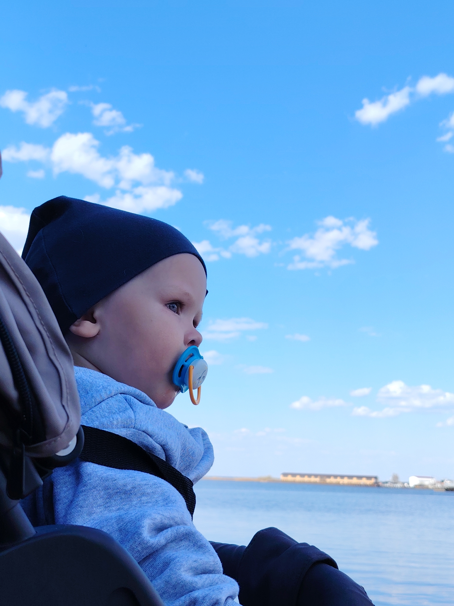 A vertical shot shows a baby sitting in a stroller with a blue pacifier in their mouth, wearing a navy blue cap and light blue hooded sweatshirt. The baby is fastened in with black straps. The stroller's frame is partially visible, a mix of gray and black.
Behind the baby is a body of water, likely a river or large lake, with a faint, indistinct shoreline in the distance. Structures are visible on the shoreline, including what appears to be a long, rectangular building.
The sky occupies the upper portion of the frame and is a bright blue with scattered white clouds.
The photo was taken near Chaikovsky, Russia.