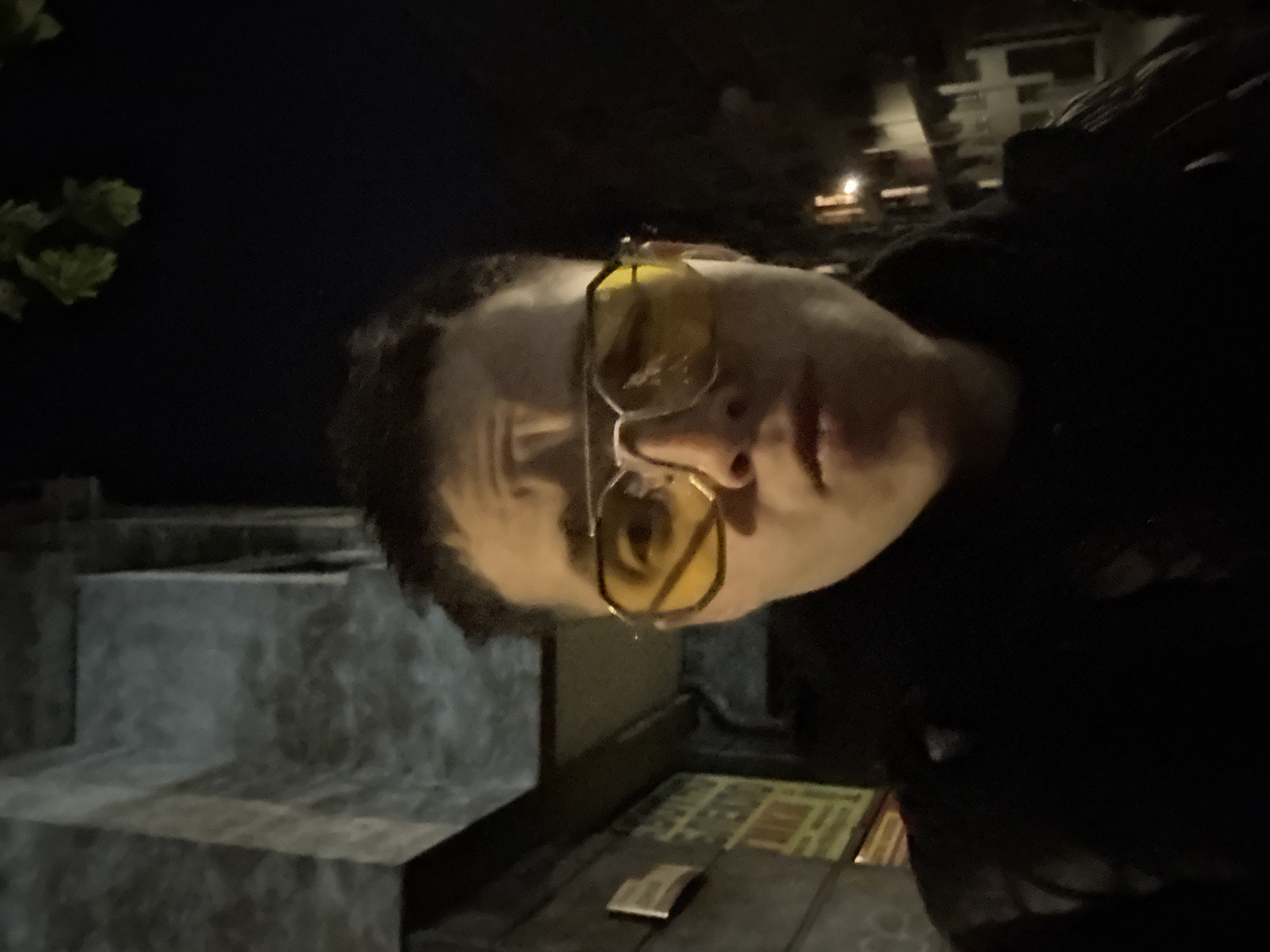 A person wearing yellow-tinted aviator-style glasses is the primary subject. They are outdoors at night. To the left, there appears to be a concrete structure, possibly stairs or a retaining wall, and foliage. The background shows dim lights indicating a city environment. Based on contextual clues and assuming the location request is accurate, the scene is possibly in Sofia, Bulgaria. The photo appears to be taken from a low angle, perhaps as a selfie.