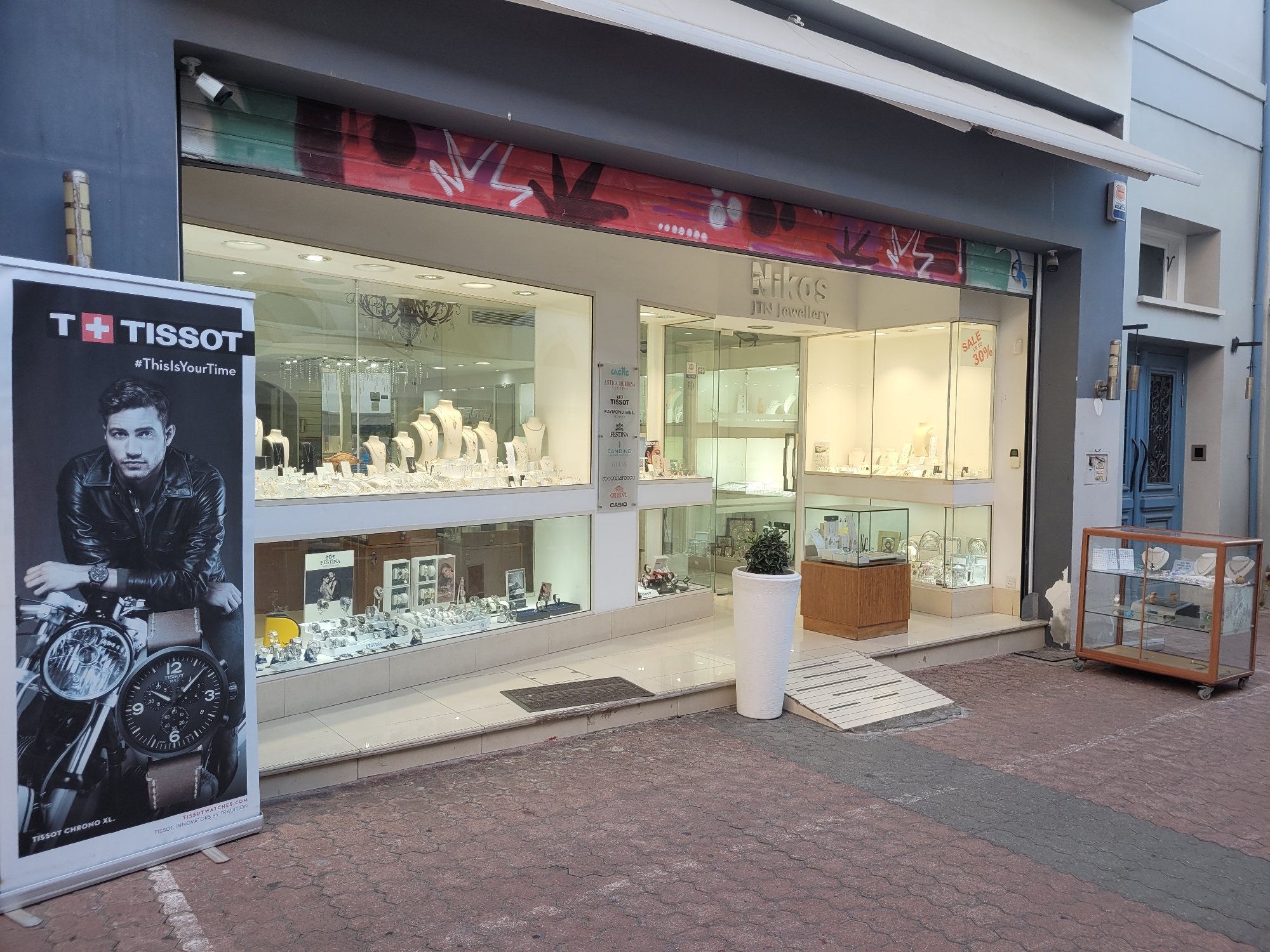 The image shows the exterior of a jewelry store named "Nikos IIN Jewellery" located in Limassol, Cyprus. A prominent Tissot advertising banner is positioned to the left of the store entrance. The store features large display windows showcasing a variety of necklaces, rings, and watches. A sign inside one of the windows indicates a "SALE UP TO 30%". The street in front of the store is paved with red and gray interlocking bricks. A white ramp leads up to the store entrance. To the right of the entrance, a glass display cabinet on wheels is situated on the sidewalk. The building facade above the store has graffiti artwork. A security camera is visible mounted on the building above the store entrance.