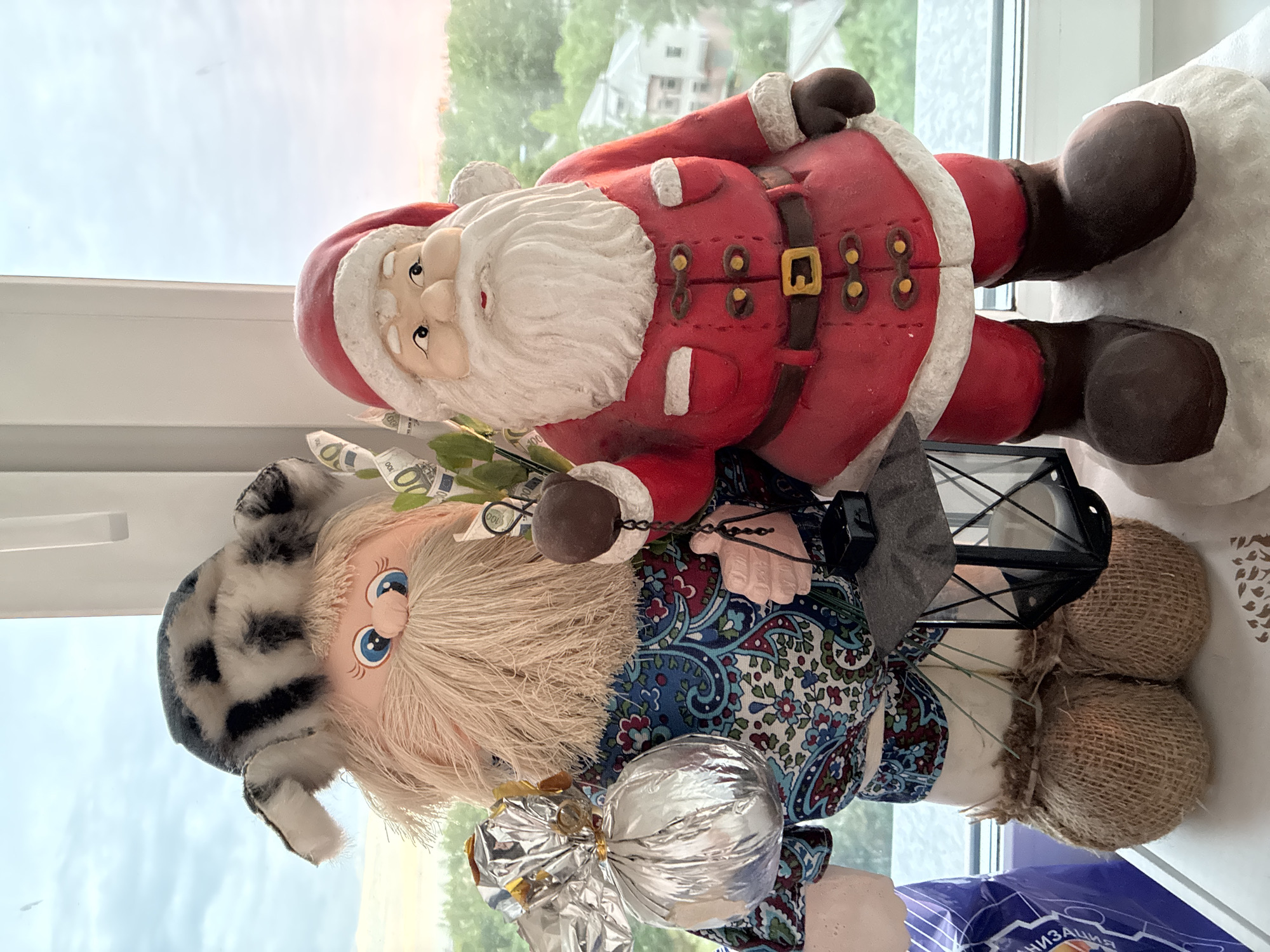 Two decorative figurines are positioned on a white windowsill, overlooking an outdoor scene. The left figure is a doll-like entity with a light-colored beard, blue eyes, and a grey and black patterned faux-fur hat. It wears a blue, red, and green paisley-patterned shirt, light-colored pants, and brown burlap-textured feet. In its left hand, it holds a silver-wrapped spherical object, and in its right hand, a dark metallic lantern. Behind its right hand, a small arrangement of green stalks decorated with miniature 100 Euro banknotes is visible. The right figure is a Santa Claus figurine, dressed in a red suit with white trim, a red hat, and brown boots, standing on a white textured base resembling snow. The outdoor background features an overcast sky with an orange glow near the horizon, indicating late afternoon or early evening. Below the horizon, green trees and foliage are present, with distant residential buildings partially visible. The location is an unknown city, unknown country.
