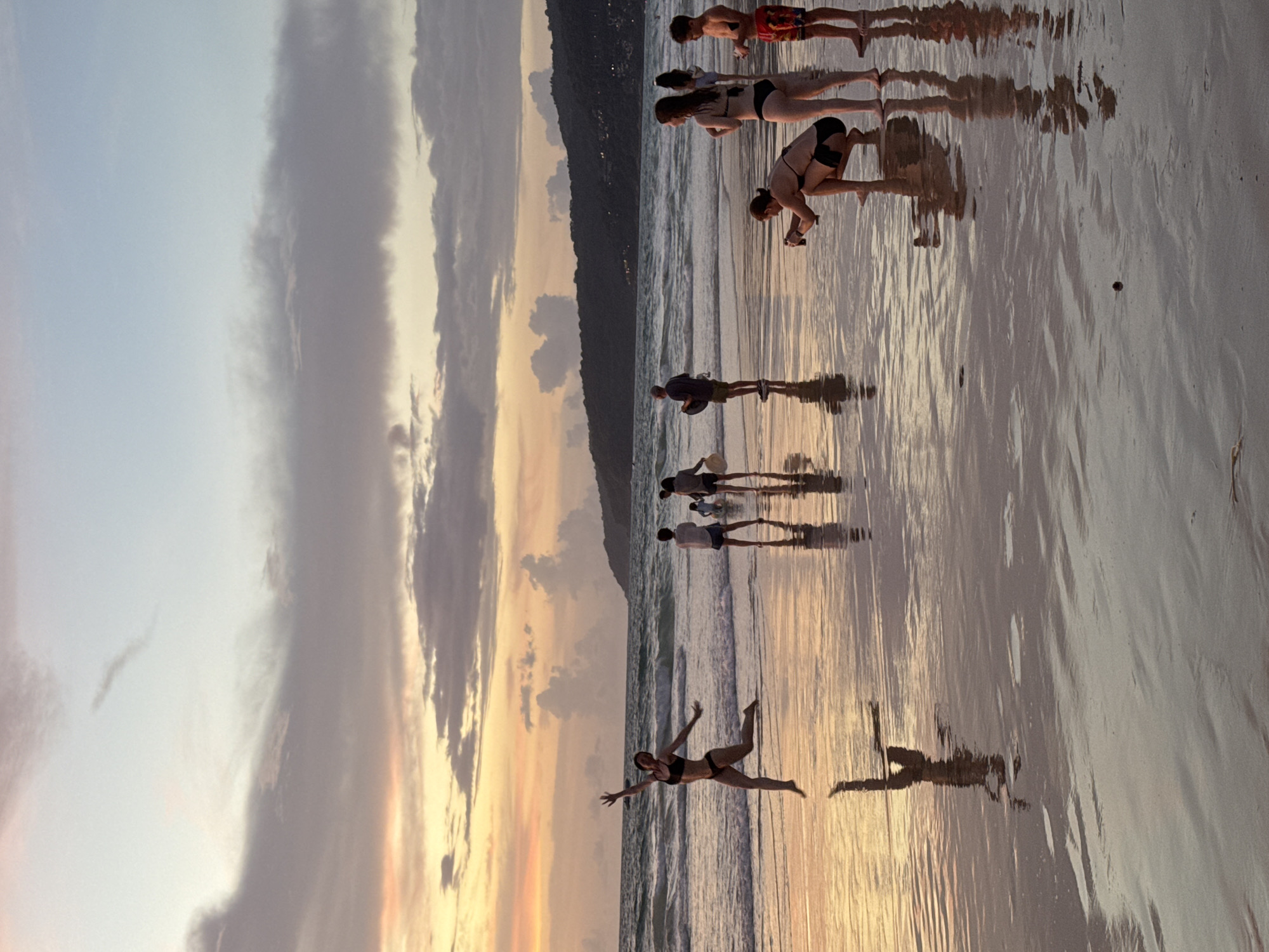 At sunset on Karon Beach, Thailand, a woman in a black bikini jumps joyfully in the shallow water. Her reflection is visible on the wet sand. In the background, several other people are walking along the shore, with a hill forming the backdrop to the scene. The sky is filled with dramatic clouds, painted in hues of orange, pink, and gray, reflecting the fading light.