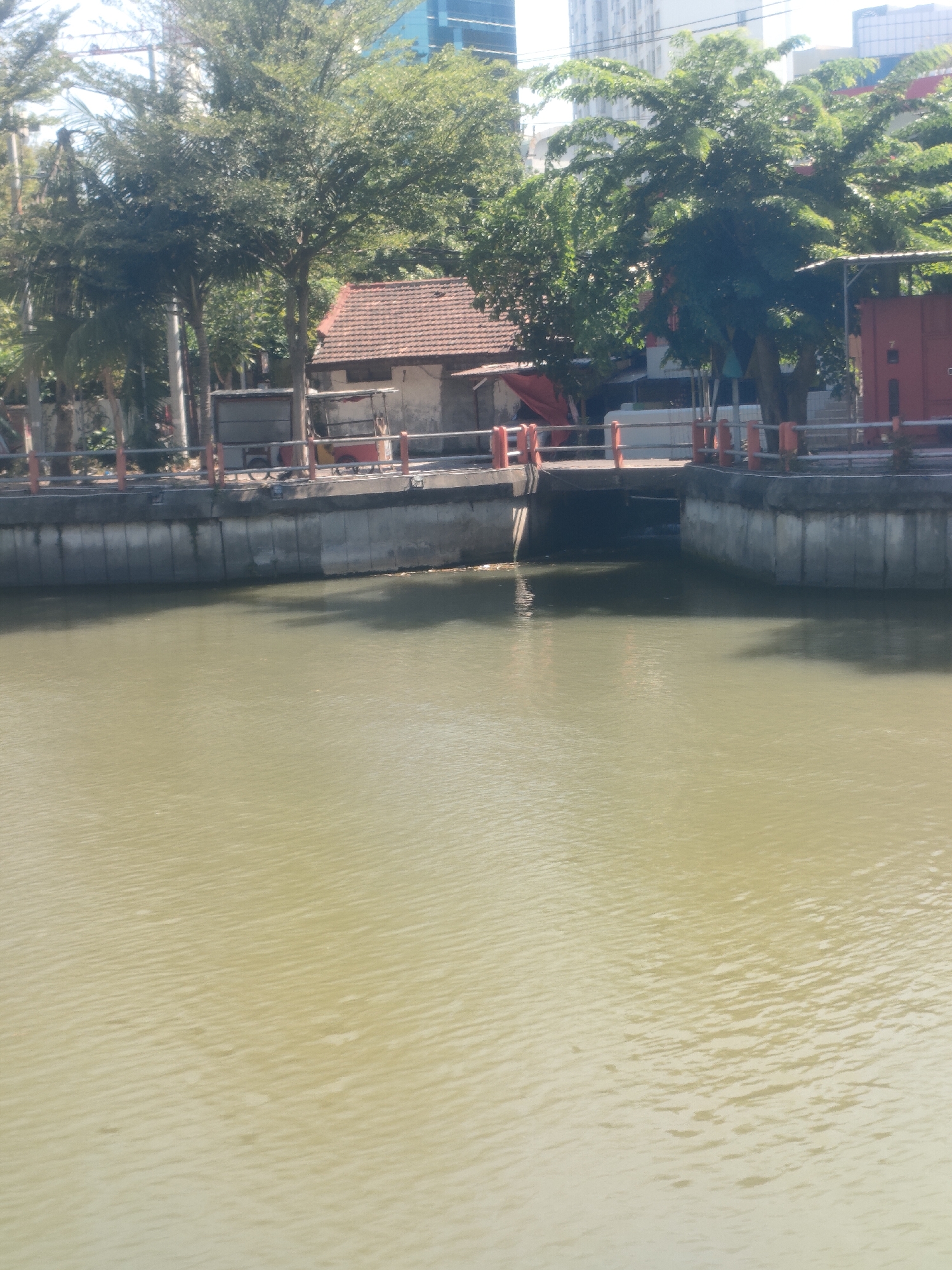 The image depicts a canal in Surabaya, Indonesia, with a bridge crossing it. On the left side of the canal, there is a concrete wall with a railing and trees in the background. A small building with a red-tiled roof is visible behind the railing. On the right side of the canal, there is a red shipping container. The canal water is murky and calm. The overall scene is urban and appears to be a functional part of the city's infrastructure.