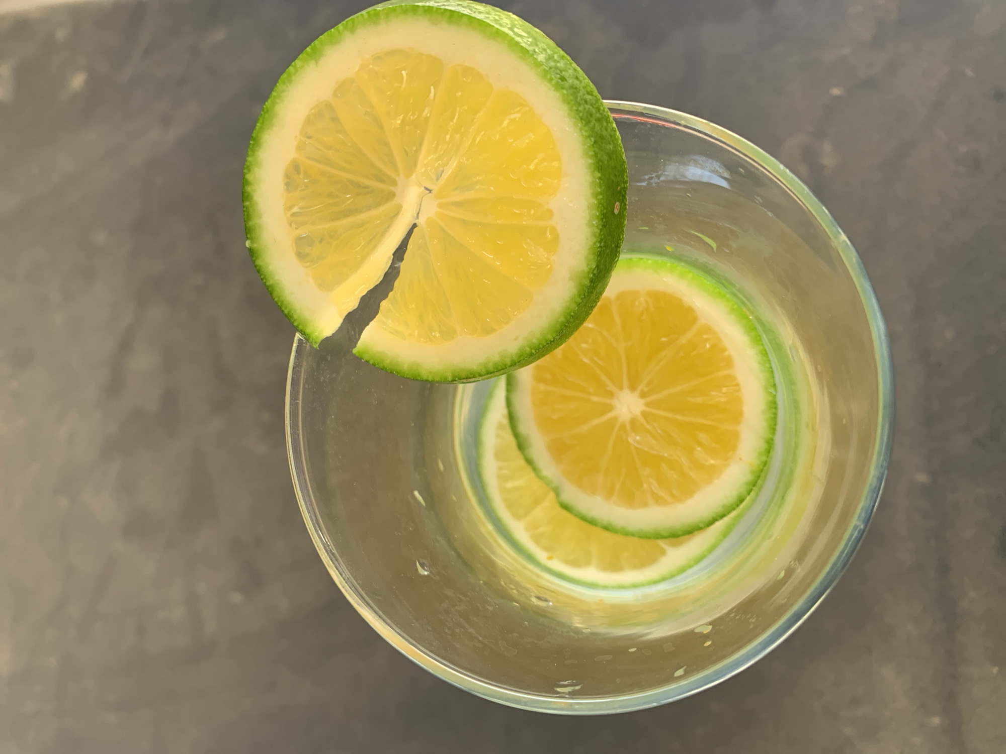 The image depicts a clear glass containing two lime slices submerged in liquid, with a third lime slice perched on the rim of the glass. The limes have a yellow interior and a green rind. The background is a grey surface. The city and country are irrelevant to the image content.