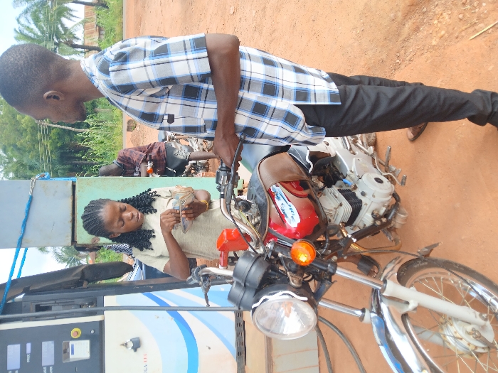 The scene depicts a fuel station in Adikpo, Nigeria, set on reddish-brown dirt ground with green foliage and distant structures under an overcast sky. In the foreground, a male subject wearing a short-sleeved blue, black, and white plaid shirt and dark trousers stands beside a red and chrome motorcycle, holding its handlebars. To the immediate left of the motorcycle, a female subject with braided dark hair, wearing a light-colored short-sleeved top, is intently counting a stack of banknotes held in her hands. A white and blue fuel dispensing pump is partially visible to the far left. A second male subject is partially visible in the mid-ground background, standing near another motorcycle to the right of the fuel pump.