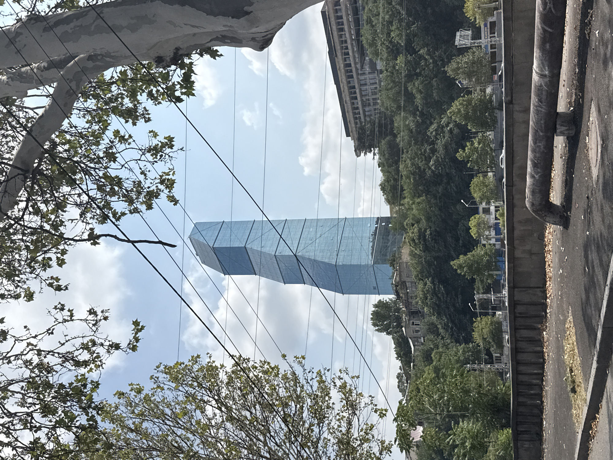 A tall, modern skyscraper with a blue glass facade dominates the upper center of the frame. It has a unique, twisted shape. In front of the skyscraper, a less modern, long building with multiple floors and balconies is visible. The buildings are situated on a hill covered with lush green trees. The sky is partly cloudy with scattered white clouds against a blue background. In the foreground, a dark, segmented concrete wall runs horizontally across the image, separating the viewer from a street or pathway. A large, weathered metal pipe is prominently featured in the lower foreground, resting on a small concrete block. Overhanging branches from trees partially obscure the top of the image, and power lines are strung across the sky. The scene appears to be an urban landscape in Tbilisi, Georgia.