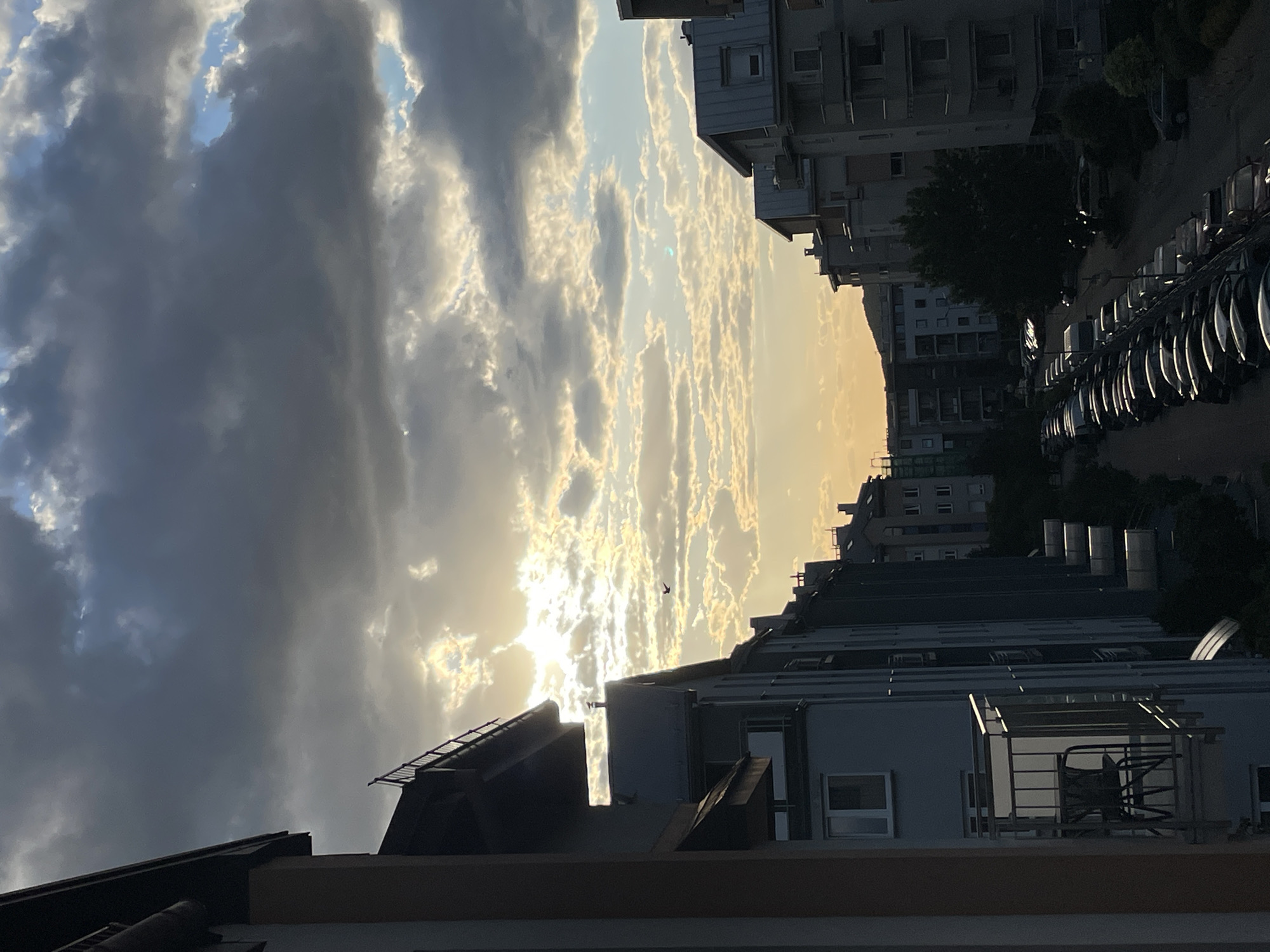 Here is a description of the image:
The image depicts an urban scene with buildings and a cloudy sky. The buildings vary in height, with some appearing to be residential apartments. A street with parked cars runs alongside the buildings. The sky is filled with clouds that range from dark and heavy to light and wispy. The sun is partially obscured by the clouds. A bird is flying in the distance against the bright sky. The image appears to be taken at dusk or dawn, given the light.