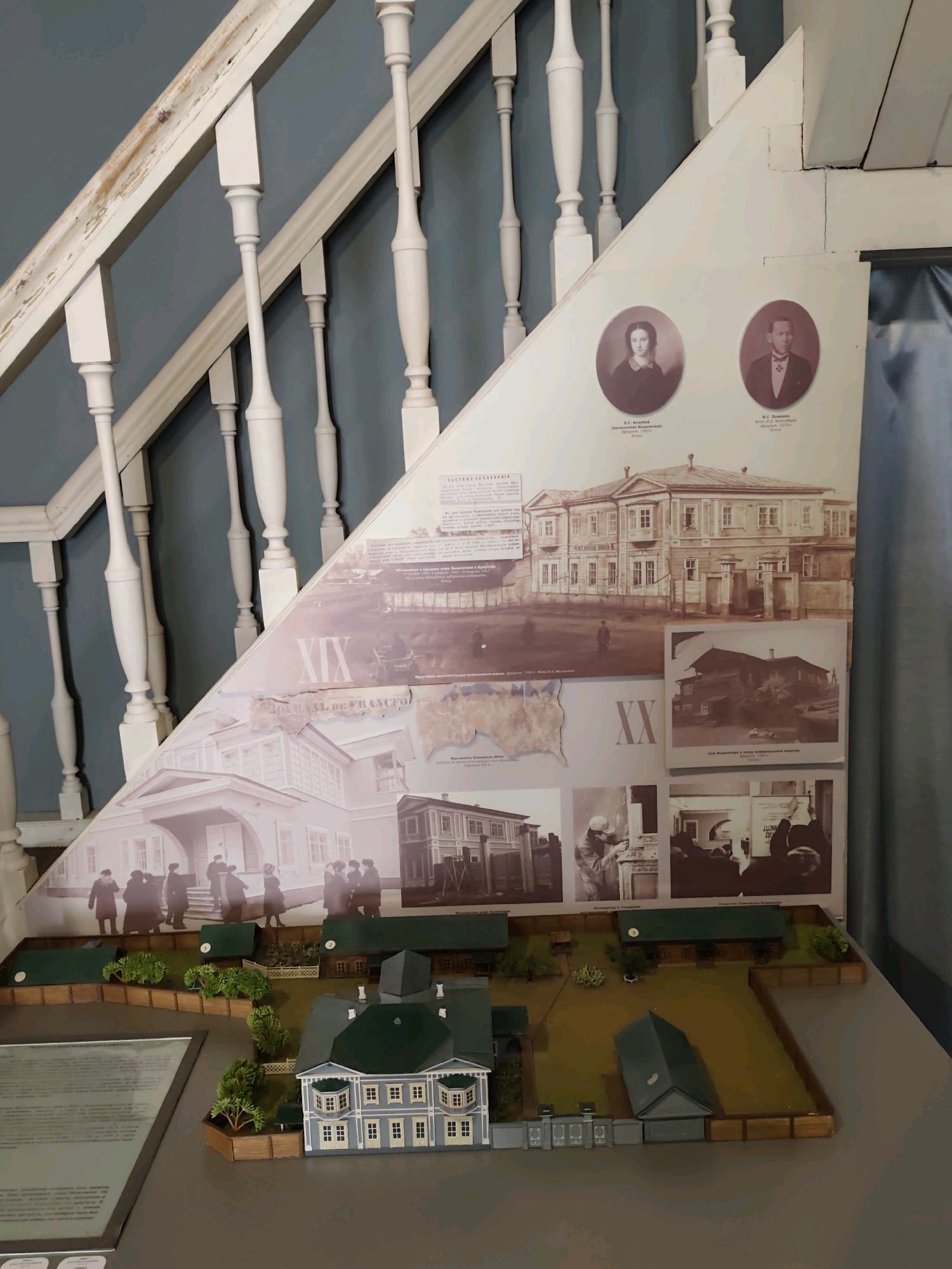 The image depicts a museum exhibit featuring a diorama of a historical estate with a large, multi-story building as the centerpiece, alongside smaller outbuildings and a landscaped garden. The diorama is positioned on a gray surface, possibly a display table. Above the diorama, a large, triangular informational display is mounted on a pale blue wall, mimicking the angle of a staircase. This display includes portraits of a man and a woman in oval frames, identified with Russian text. Below the portraits, various historical images are collaged, showing architectural renderings of buildings, street scenes with figures, and what appears to be a map. The overall impression is a historical exhibit focused on a specific location. The Russian text on the display, when deciphered with additional information, indicates this exhibit relates to the city of Irkutsk, Russia, and specifically to the estate of the Trubetskoy family, a prominent Decembrist family who were exiled to Irkutsk. The exhibit aims to showcase the historical significance and architectural aspects of their former residence.
