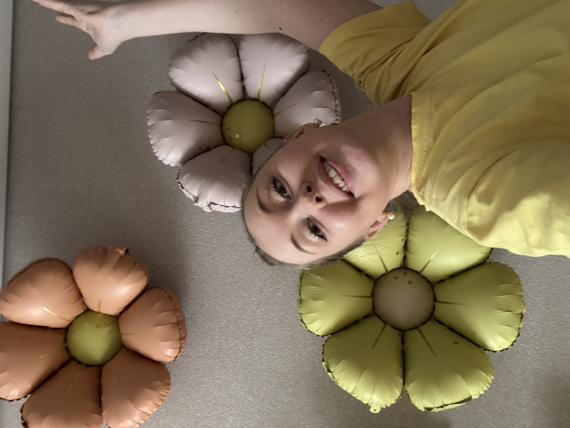 A fair-skinned person with visible teeth is lying on the floor next to three flower-shaped balloons. One balloon is blush pink, one is peach, and one is lime green, and each has a green center. The person is wearing a yellow shirt. The location is Unknown, Unknown.