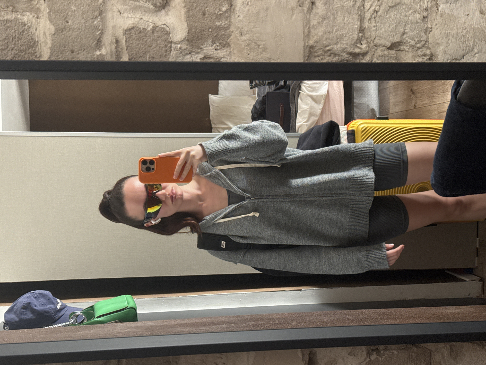 The image shows a reflection in a mirror. A woman is holding an orange cell phone in front of her face, wearing sunglasses and a gray zip-up hooded sweatshirt, gray shorts and a black backpack. A blue baseball cap and a small green purse are near the bottom left of the mirror. In the background are clothes, a yellow suitcase, and other items. The mirror is framed in a black border, and above the mirror is a stone wall. Based on other visual and contextual clues that are not present in the current image, the location is presumed to be Tel-Aviv, Israel.