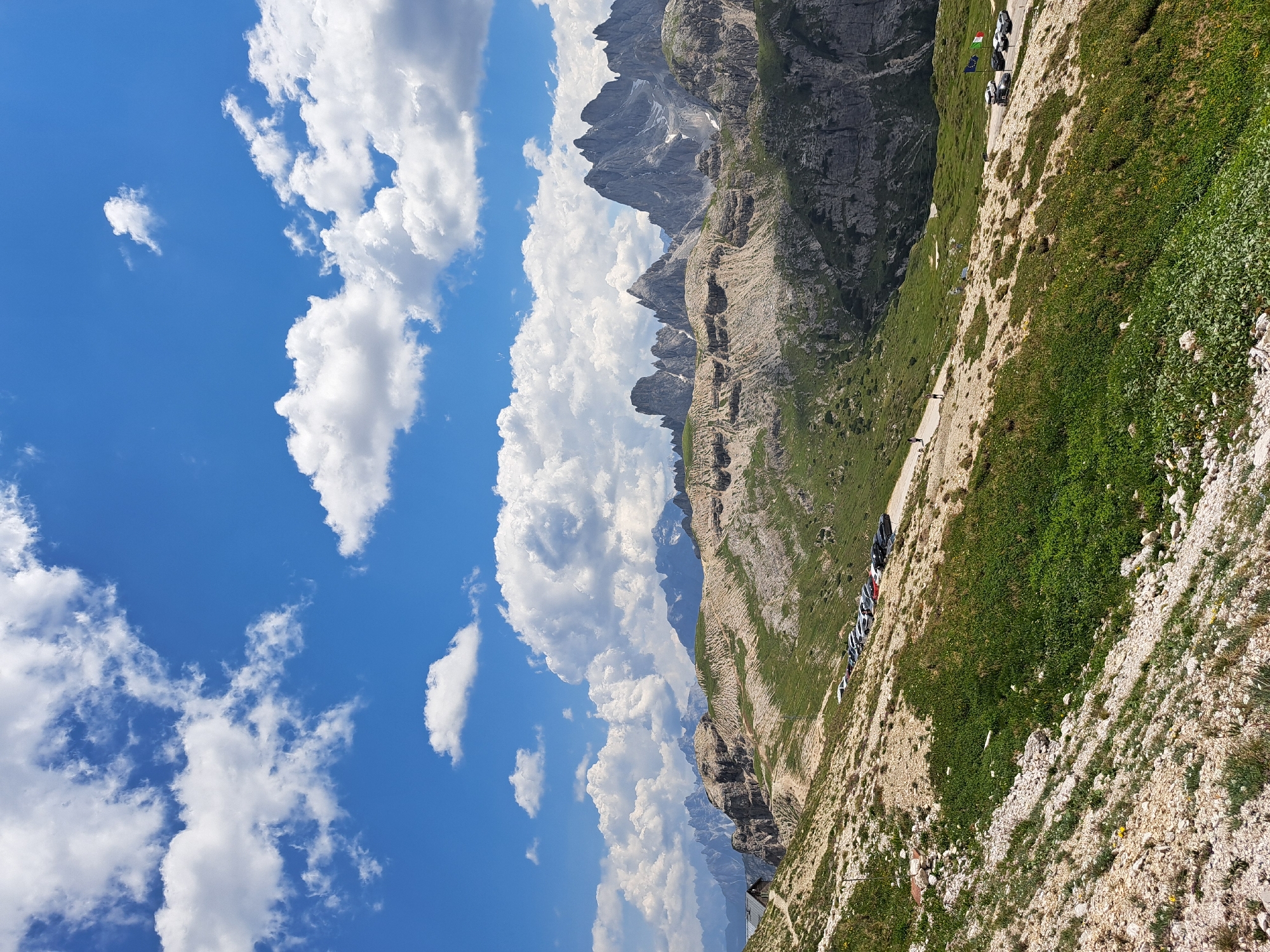 The image depicts a mountain landscape with a clear blue sky dotted with fluffy white clouds. The foreground is a steep, rocky slope covered in sparse green vegetation and small yellow flowers. A gravel path runs along the side of the slope, bordered by a low stone wall.
Several cars are parked along the side of the path, and in the distance, there are glimpses of people walking. The background features jagged, rocky mountain peaks, some with patches of snow, under the bright daylight. The overall scene suggests a hiking or scenic viewpoint in a mountainous region. The sun appears to be high in the sky, indicating it is likely daytime. The weather appears to be fair and sunny. The Italian flag is visible near the parked cars, hinting at the location potentially being in Italy. There is no discernible text within the image.