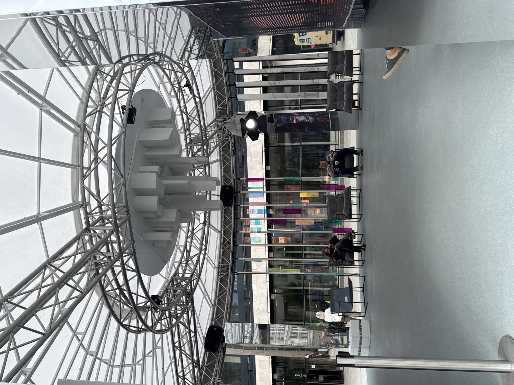 The image depicts an outdoor public space with a large, modern, circular canopy structure overhead. The canopy is supported by a complex network of metal trusses and features a translucent white material. Hanging from the center of the canopy is a multi-tiered cylindrical structure with rectangular protrusions.
In the foreground, the ground is a smooth, light gray surface. To the left, a silver metal stanchion with a rounded base stands vertically. Two white boxes with Cyrillic text are visible behind the stanchion.
Further back, a group of dark gray lounge chairs are arranged on the gray ground. In the center-left, two individuals are seated in chairs. One person, facing away from the camera, is looking down at their phone. Another person, also facing away, is seated.
To the right of these chairs, more identical dark gray lounge chairs are present. In the center-right, a person is seated in a chair, looking down at their phone. Further to the right, the scene is dominated by highly reflective, mirrored surfaces that surround the area, reflecting the canopy structure and the surrounding buildings.
Behind the seating area, a row of buildings with large glass windows is visible. Several colorful, rectangular elements hang vertically from the canopy structure in front of the buildings. Two black speakers are mounted on the metal structure of the canopy.
On the far right side of the image, a large digital screen displays a pixelated image of a cityscape. A yellow vehicle is parked in the background to the right of the buildings. The overall environment suggests a contemporary urban setting, possibly an exhibition space or public plaza in Moscow, Russia, given the Cyrillic text and the architectural style.