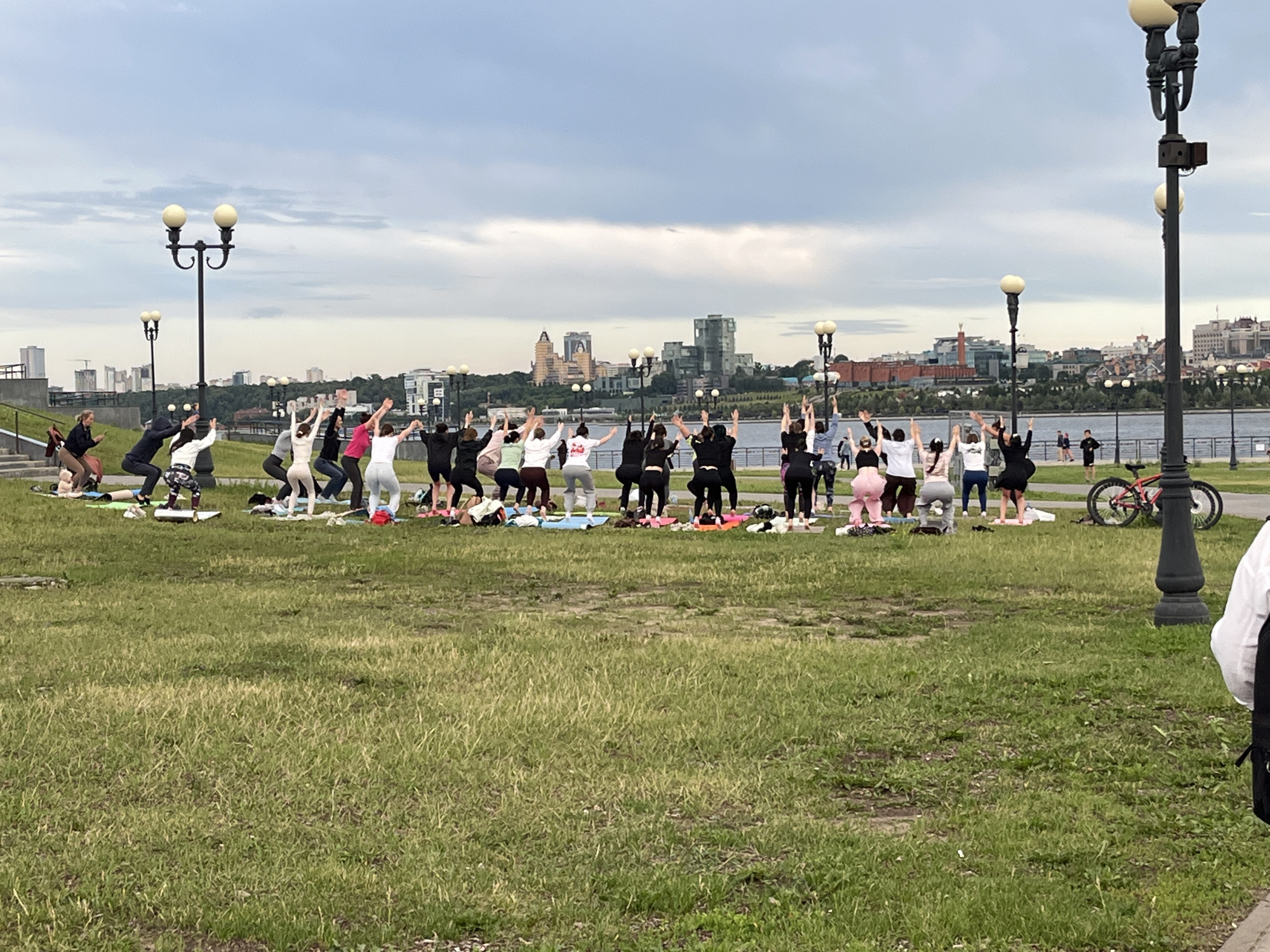The image shows a group of people practicing yoga on mats on a grassy area. The group is facing the right side of the frame. The people are wearing athletic clothing. In the background, there is a body of water with a city skyline on the other side. The city skyline includes various buildings, some of which appear to be historical. There are also lampposts along the waterfront. A red bicycle is parked on the right side of the frame. The sky is cloudy. The image appears to be taken in an outdoor public space, likely a park or waterfront promenade. The question implies this location is in Kazan, Russia.