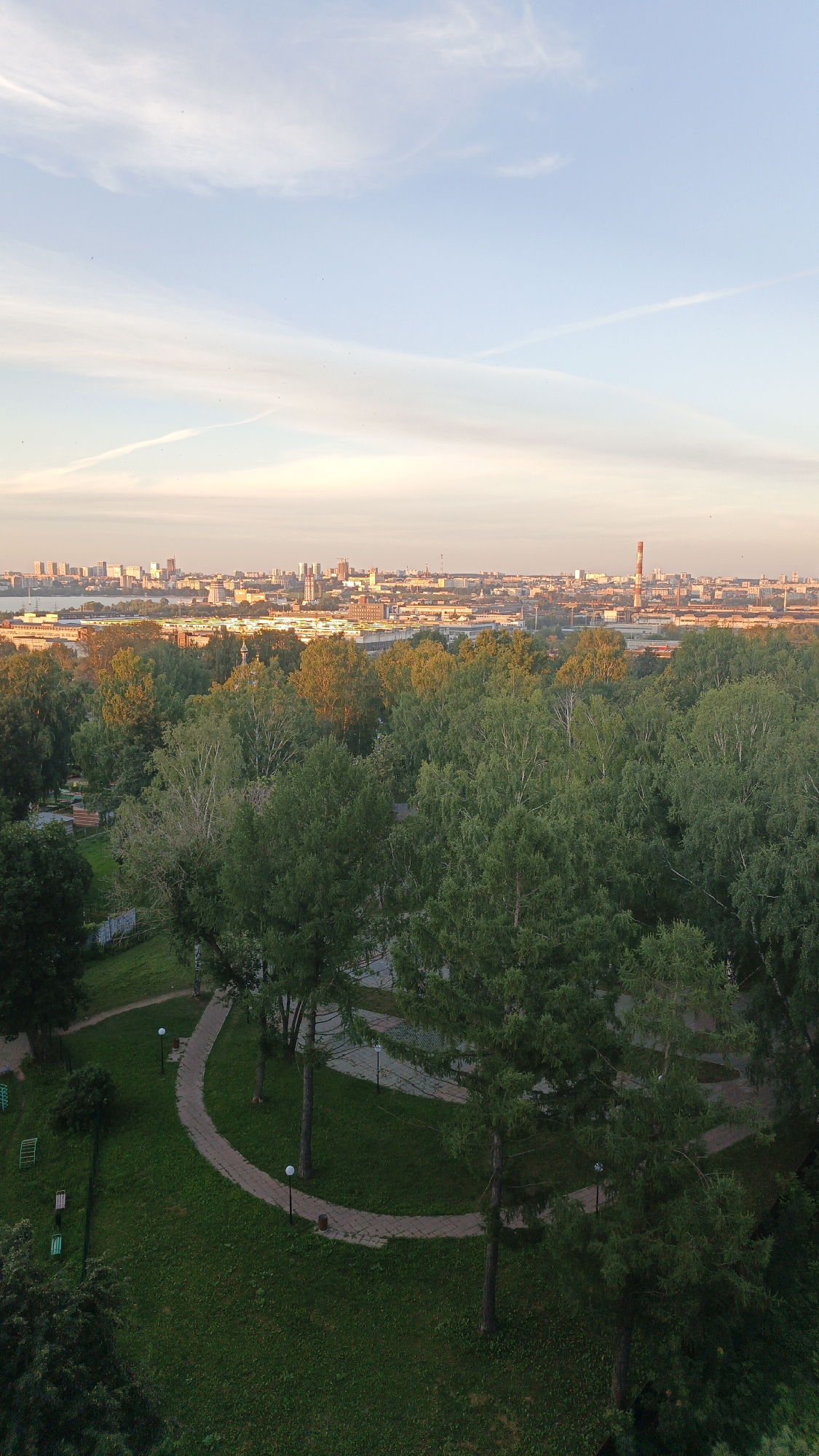 This aerial, full-frame, outdoor, vertical shot captures a city skyline and park on a clear day. The sky is a pale blue with wispy white clouds and faint contrails. Below the sky, a cityscape stretches across the horizon, featuring numerous buildings of varying heights and styles. A prominent industrial chimney, painted red and white, stands out among the structures. In the foreground, a lush green park area dominates the view. Winding pathways, lit by lampposts, meander through the grassy expanse. Tall, mature trees with dense green foliage are scattered throughout the park, with some closer trees casting shadows on the ground. The park appears to be part of a residential or public area, with some benches visible in the lower left corner. The lighting suggests it might be late afternoon or early morning, with the sun casting a warm glow on the distant buildings. The overall scene depicts a harmonious blend of urban development and natural greenery. The provided information states this is a view of Izhevsk, Russia.