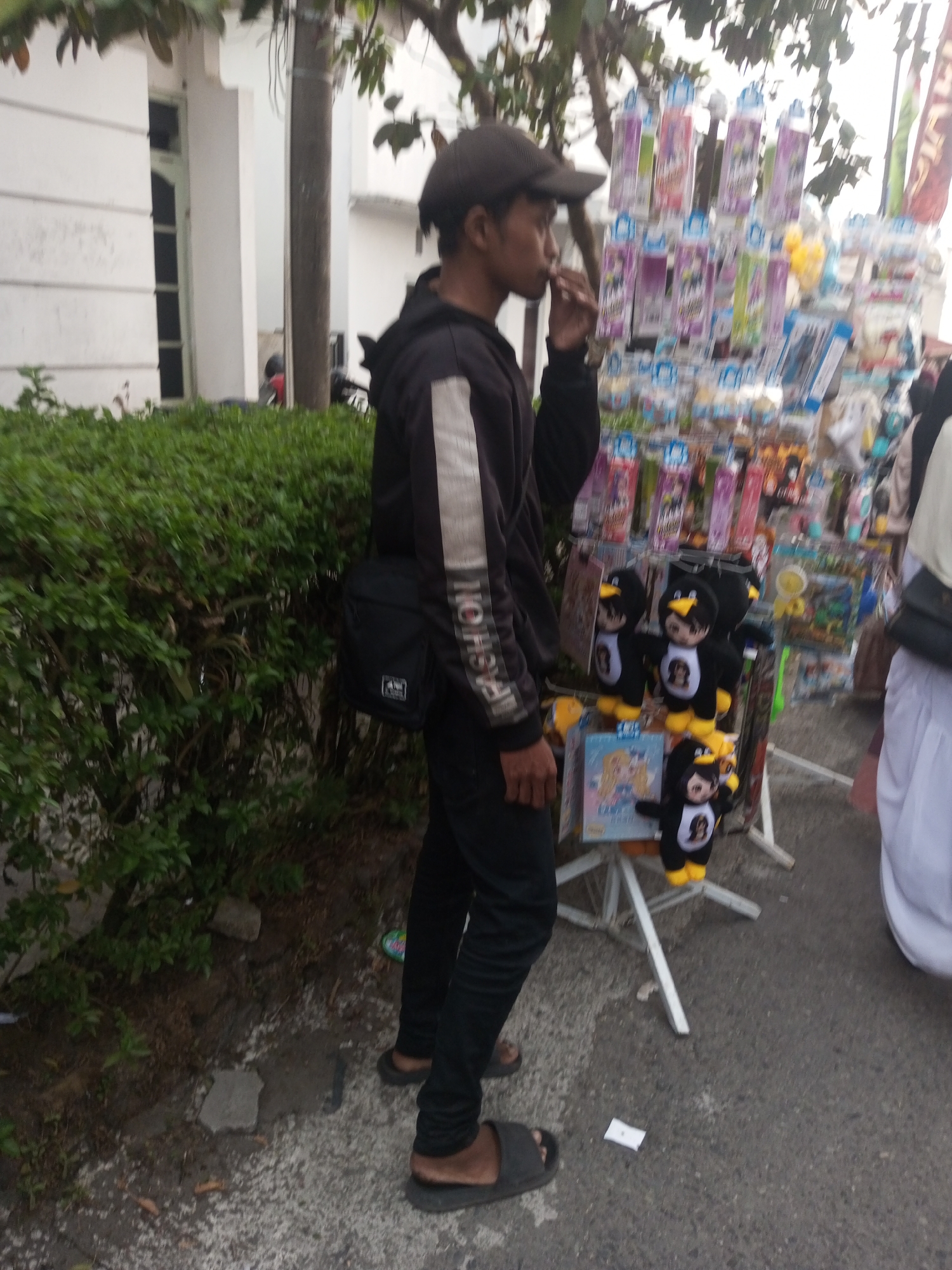 A young man stands on a paved surface, facing right, with his left hand near his mouth. He is wearing a dark baseball cap, a black hooded jacket with white stripes on the sleeves, and black pants. A black crossbody bag is slung over his left shoulder. Beside him, a display rack filled with various items, including plush toys resembling penguins, is visible. In the background, a white building with windows and some greenery can be seen. The scene appears to be outdoors in an urban environment.