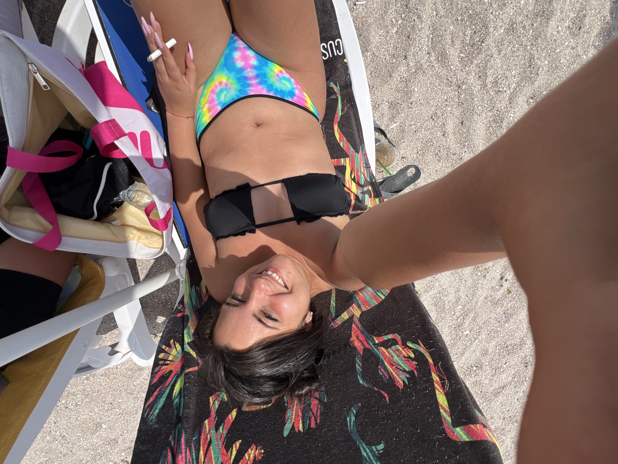 A young woman with dark hair and a tan is lying on a beach chair on a sandy beach in Pomorie, Bulgaria. She is wearing a black bikini top and a colorful tie-dye bikini bottom. She is holding a lit cigarette in her right hand. In the foreground, a white beach bag with pink straps is visible. The woman is smiling and looking at the camera. The background is a sandy beach with a few scattered seashells.