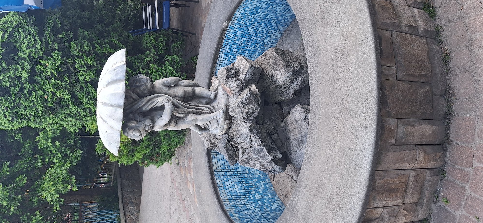 The image shows a stone fountain with a statue of two cherubs holding an umbrella. The cherubs are made of light-colored stone and are standing on a cluster of large rocks in the center of the fountain. The fountain itself is circular with a concrete rim and a blue tiled interior. The fountain is surrounded by a stone wall, and behind it are trees and foliage. In the background, there are some blue chairs and a fence. The overall impression is that of an outdoor public space, possibly a park or a courtyard. The image was taken in Bila Tserkva, Ukraine.