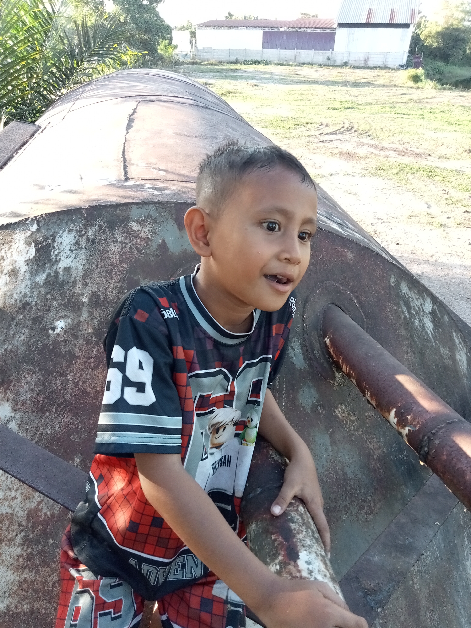 A young boy, appearing to be around five to seven years old, stands leaning against a large, weathered, cylindrical metal object, possibly a water tank or silo, in an outdoor environment. The boy is wearing a black and red checkered shirt with white and gray accents and the number "69" printed on the front. His shorts are also patterned with red and black checks and feature the number "69" as well. He has short, dark hair and dark eyes. His mouth is slightly open, and he is looking to his right. The metal structure he is leaning on is rusted and has visible seams. A thick, rusty pipe protrudes from the side of the cylindrical object.
The background shows a grassy area with scattered dirt patches. In the distance, there are two buildings with white and corrugated metal roofs. To the left of the frame, there are palm fronds, indicating a tropical or subtropical location. The lighting suggests it is daytime, with sunlight casting shadows. The overall scene appears to be in a rural or semi-rural setting. The location is identified as Kualu, Indonesia.