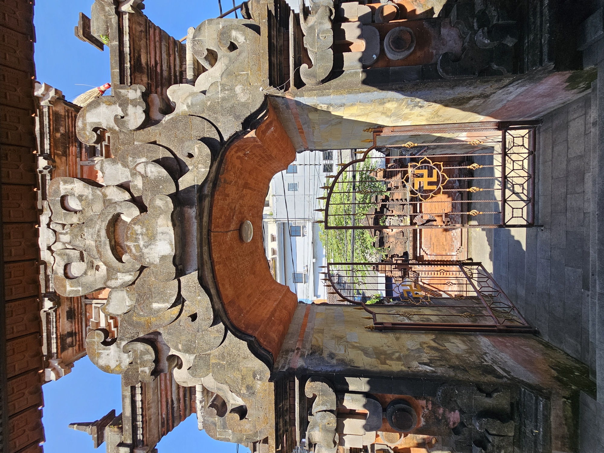 The image captures a detailed view of a traditional Balinese temple entrance, likely located in Denpasar, Indonesia. The main subject is an ornate stone gateway, characterized by intricate carvings and a reddish-orange brick archway. The stonework appears weathered, showing signs of age and exposure to the elements. To the right of the entrance, a metal gate with decorative elements, including a golden swastika symbol, is ajar, revealing a courtyard beyond. This courtyard features more brick structures and greenery. The overall environment is bright and sunny, indicated by the strong shadows cast on the stonework and pavement. The background shows modern buildings, suggesting the temple is situated within an urban area. The image is taken from a low angle, looking upwards towards the gateway. No people are visible in the scene.