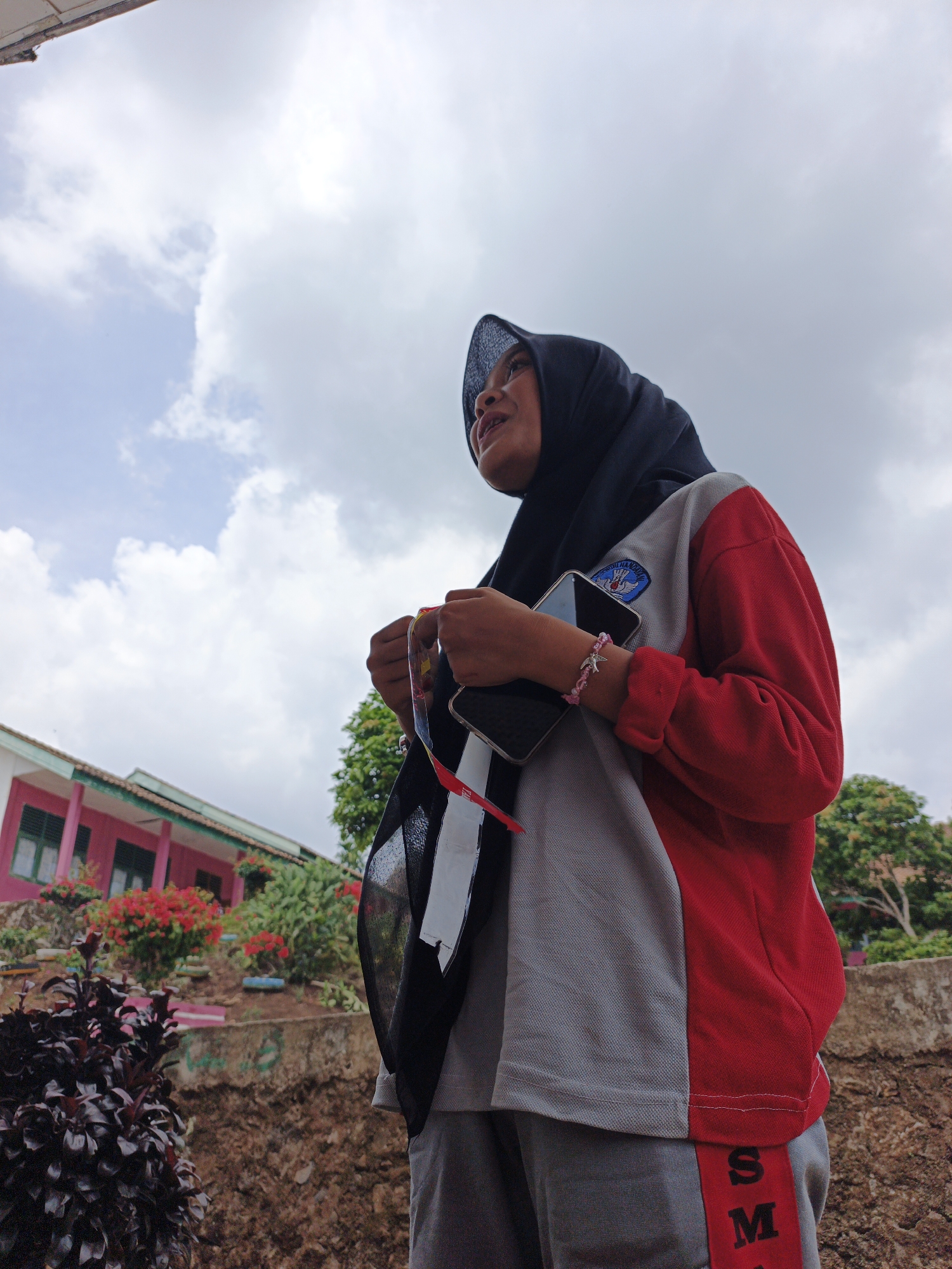 The image shows a young person wearing a black hijab and a color-blocked shirt with red and gray sections. The shirt has a patch with a blue crest and a white bird emblem. They are holding a smartphone in their right hand and a rosary with red and white beads in their left hand. A pink and silver bracelet with a star charm is visible on their left wrist. The person is looking upwards and to the left, with a slight smile. In the background, there is a pink building with green-trimmed windows, surrounded by lush greenery and red flowers. The ground is covered with brown soil and some dark purple foliage is visible in the foreground. The sky is overcast with white clouds. The scene appears to be in an outdoor setting, possibly a school campus or residential area in Jepara, Indonesia.