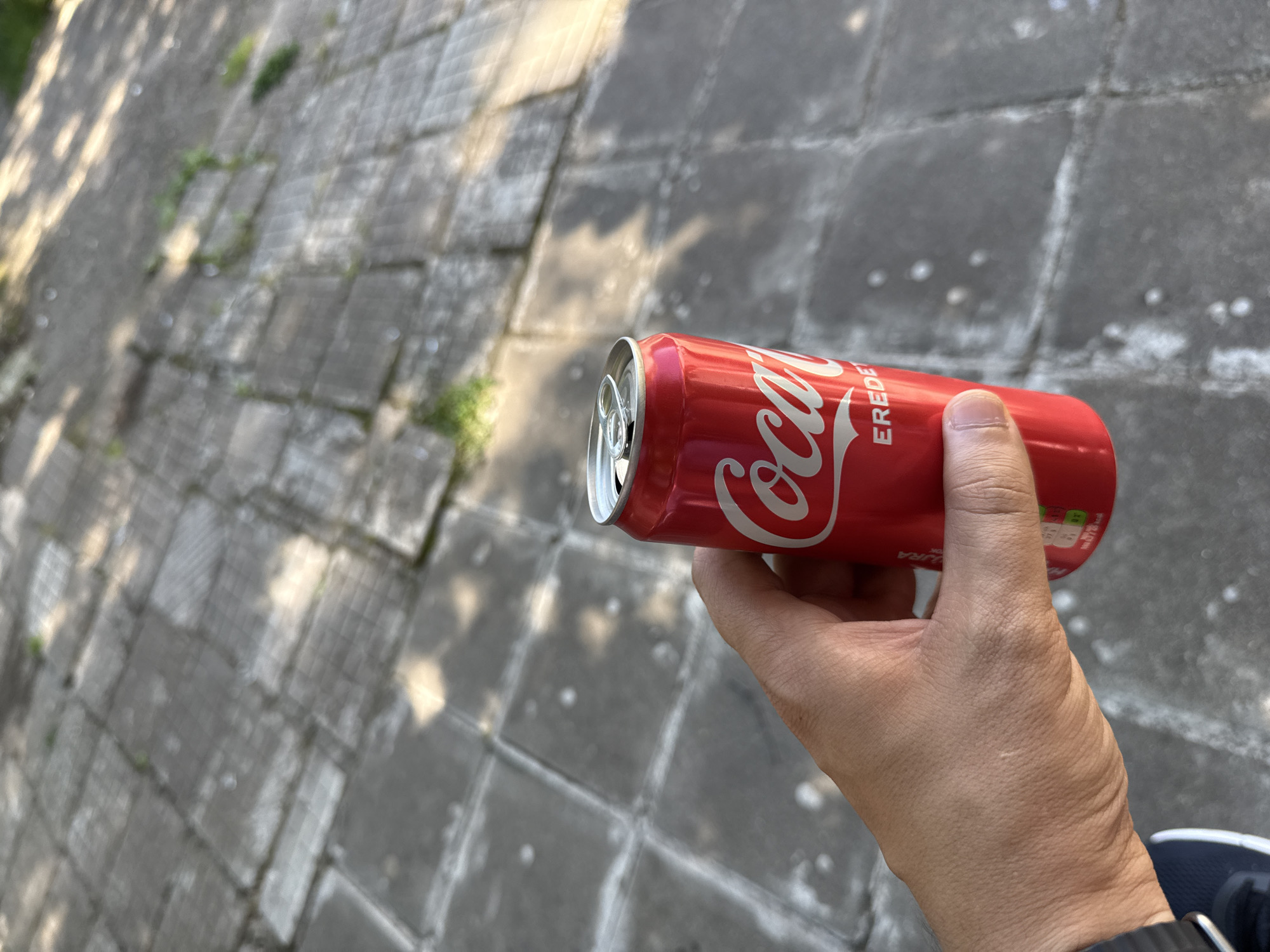 A person's hand is holding a red Coca-Cola can. The can has the Coca-Cola logo in white script and the word "EREDET" below it. The background is a grey paved surface, possibly a sidewalk or plaza, with some patches of grass growing between the paving stones. Sunlight is casting shadows on the ground, suggesting it is daytime. The scene is likely outdoors, in a public area within the city of Vratsa, Bulgaria.