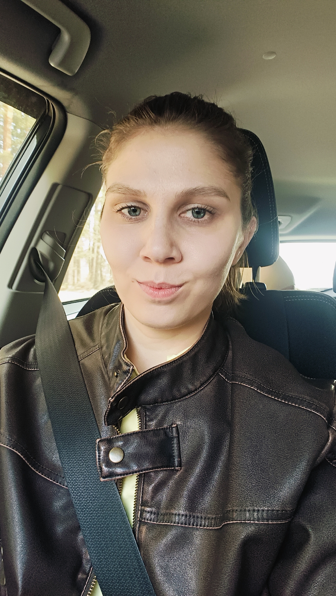 A young woman is captured in a close-up portrait, seated inside a car, likely in the passenger seat. She has light skin, blue-green eyes, and light brown hair pulled back from her face. Her expression is neutral with a subtle, slight smile or smirk, and she looks directly into the camera.
She is wearing a dark brown, possibly faux leather, jacket over a light yellow or pale green shirt. A black seatbelt is prominently draped across her chest, extending from her left shoulder towards her right hip.
The car's interior is visible, including a gray overhead lining, a black headrest behind her, and parts of the car door frame and an overhead grab handle on the left. Through the window on her left, an outdoor environment with trees and foliage can be seen, suggesting a wooded or rural setting. The lighting appears to be natural and bright, indicating it is daytime. There is no visible text in the image.