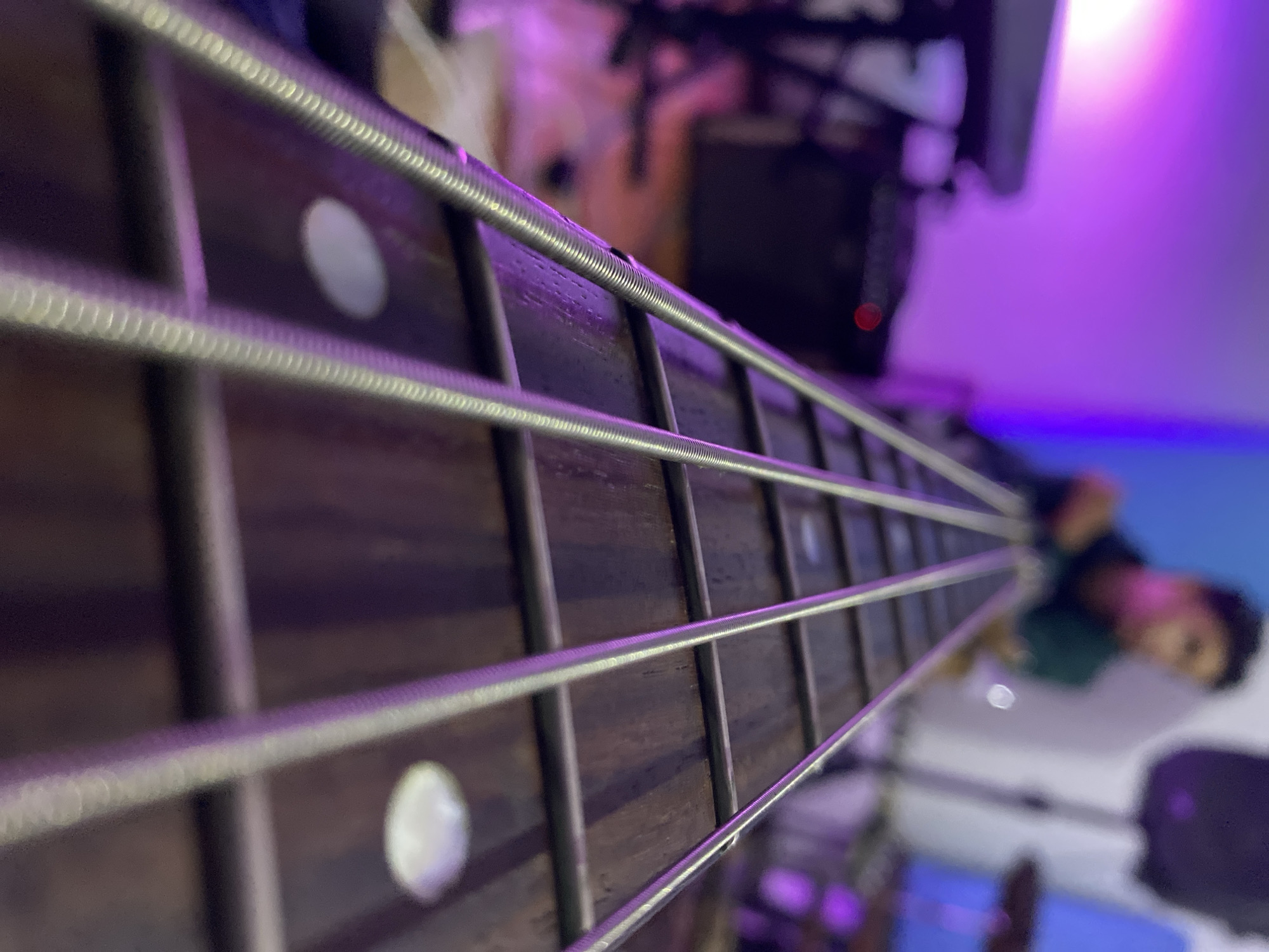 The image shows a close-up of a bass guitar neck with metal strings and fret markers. The background features blurred elements, including a person and some equipment, under purple lighting.
Location: Could not be determined, so the city of Tinaquillo, Venezuela is not confirmed.