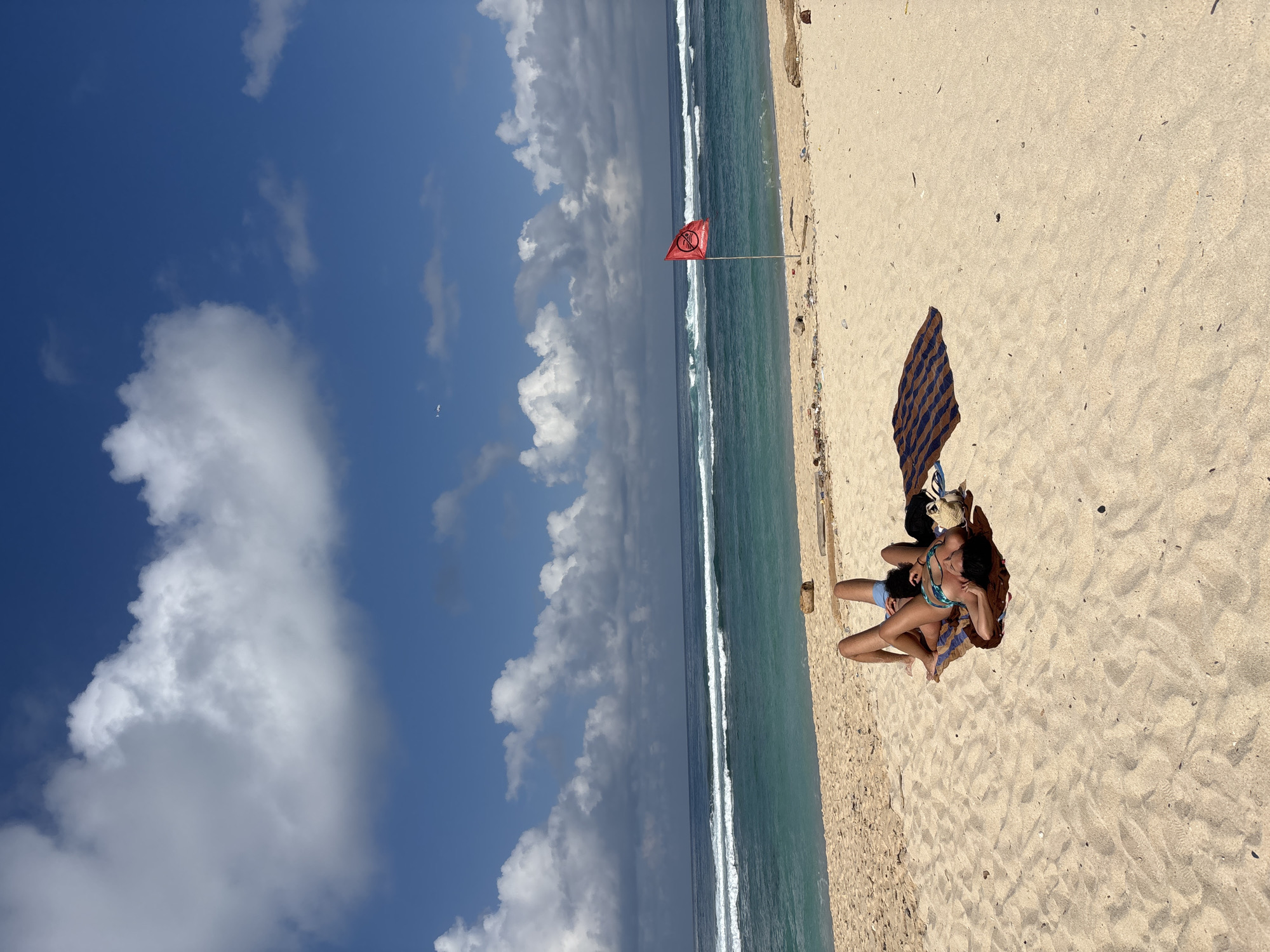 This image depicts a vibrant beach scene in Kuta Selatan, Indonesia, on a clear, sunny day. The sky, a bright blue, is dotted with scattered white cumulus clouds, some large and puffy, others smaller. Below the sky is the turquoise ocean, displaying gentle breaking waves with white foam as they roll towards the shore. The water transitions to a deeper blue further out.
The foreground is dominated by light golden sand, appearing somewhat coarse and showing minimal debris. Two individuals are relaxing on the sand towards the right side of the frame. One person, wearing a patterned swimsuit, is seen lying down, resting their head on the lap or shoulder of the other, partially visible person who is wearing light blue bottoms. They are positioned on a blue and brown striped blanket or towel, with a straw bag or hat placed nearby. They appear to be in a state of relaxation, possibly sunbathing or enjoying a quiet moment together.
A notable object in the middle distance is a red flag planted in the sand near the water's edge. It features a white circular symbol with a diagonal line through it, commonly indicating "no entry" or "prohibited," likely signifying a warning or specific zone on the beach. The bright sunlight suggests it is daytime, probably mid-morning or afternoon, and the weather is pleasant with clear skies and some clouds. The overall scene conveys a serene and tranquil atmosphere typical of a tropical beach.