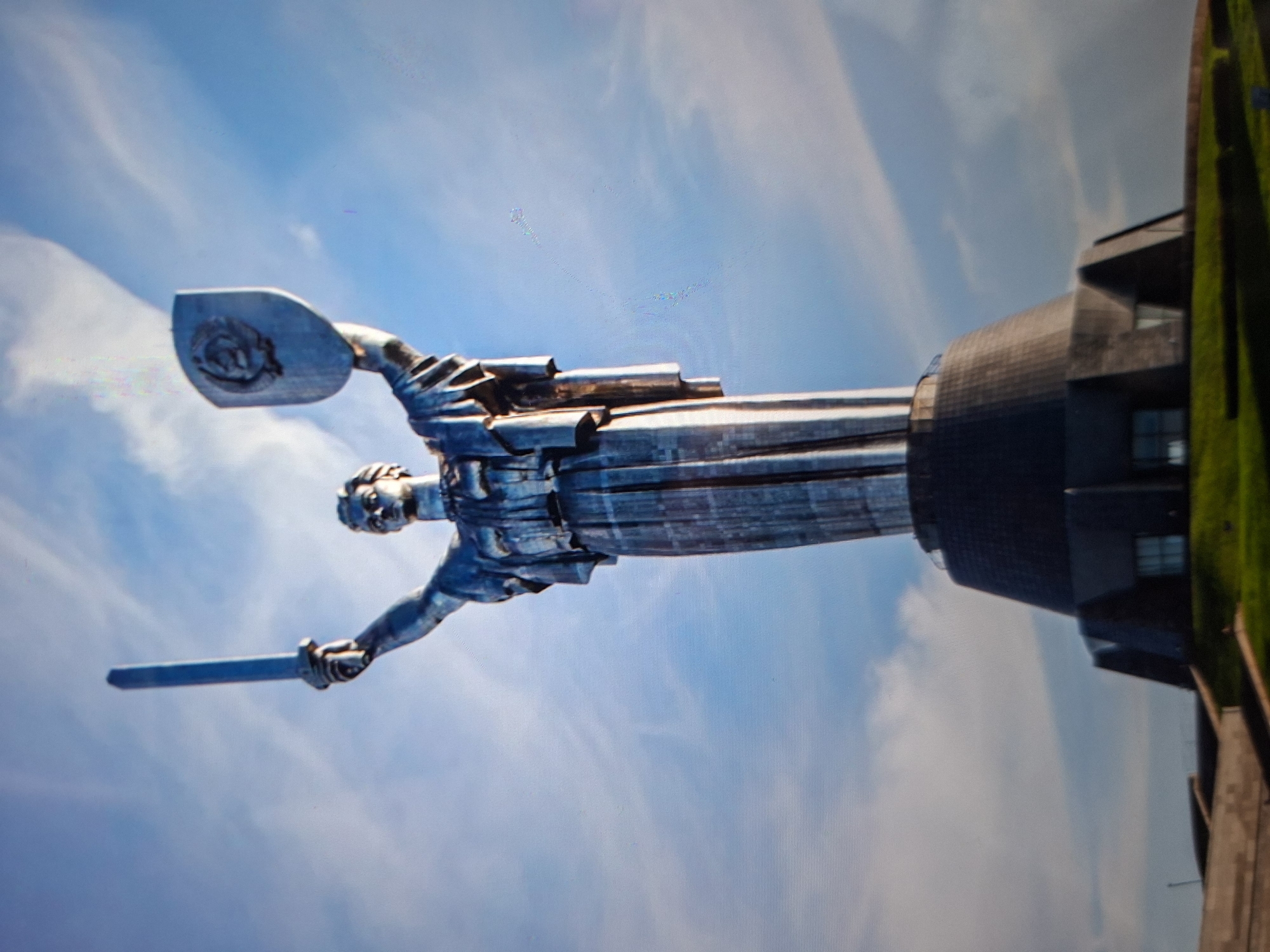 The image captures a low-angle view of the Motherland Monument in Kyiv, Ukraine, against a backdrop of a partially cloudy blue sky. The monumental stainless steel statue depicts a female figure holding a sword in her raised right hand and a shield emblazoned with the Soviet hammer and sickle emblem in her raised left hand. The statue stands atop a large, multi-tiered concrete base which is surrounded by a grassy slope. The base features a dark, angular architectural design. Sunlight illuminates the statue, highlighting its metallic surface and creating reflections. The overall scene conveys a sense of grandeur and historical significance.