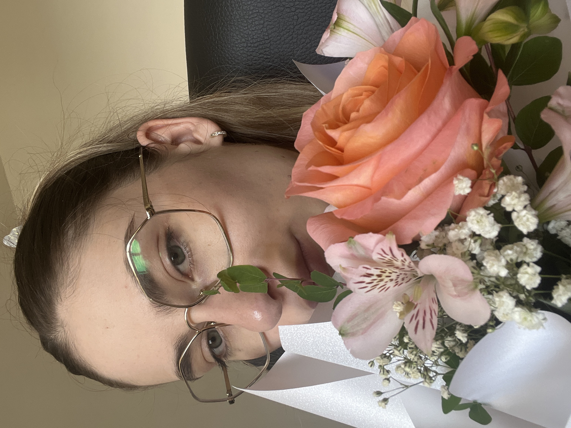 In this close-up, medium shot, a young woman wearing gold-rimmed glasses looks directly at the camera, her face partially obscured by a bouquet of flowers. The bouquet features a large peach-colored rose, pink alstroemeria with dark speckles, and small white baby's breath flowers, all wrapped in white paper. A delicate green stem with small leaves extends upwards between the woman's face and the flowers. Her hair is blonde and pulled back. She is wearing a small, sparkly hoop earring in her visible ear. The background is a plain, light-colored wall, and a dark leather chair is visible behind her. The lighting suggests an indoor setting, possibly a home or office.