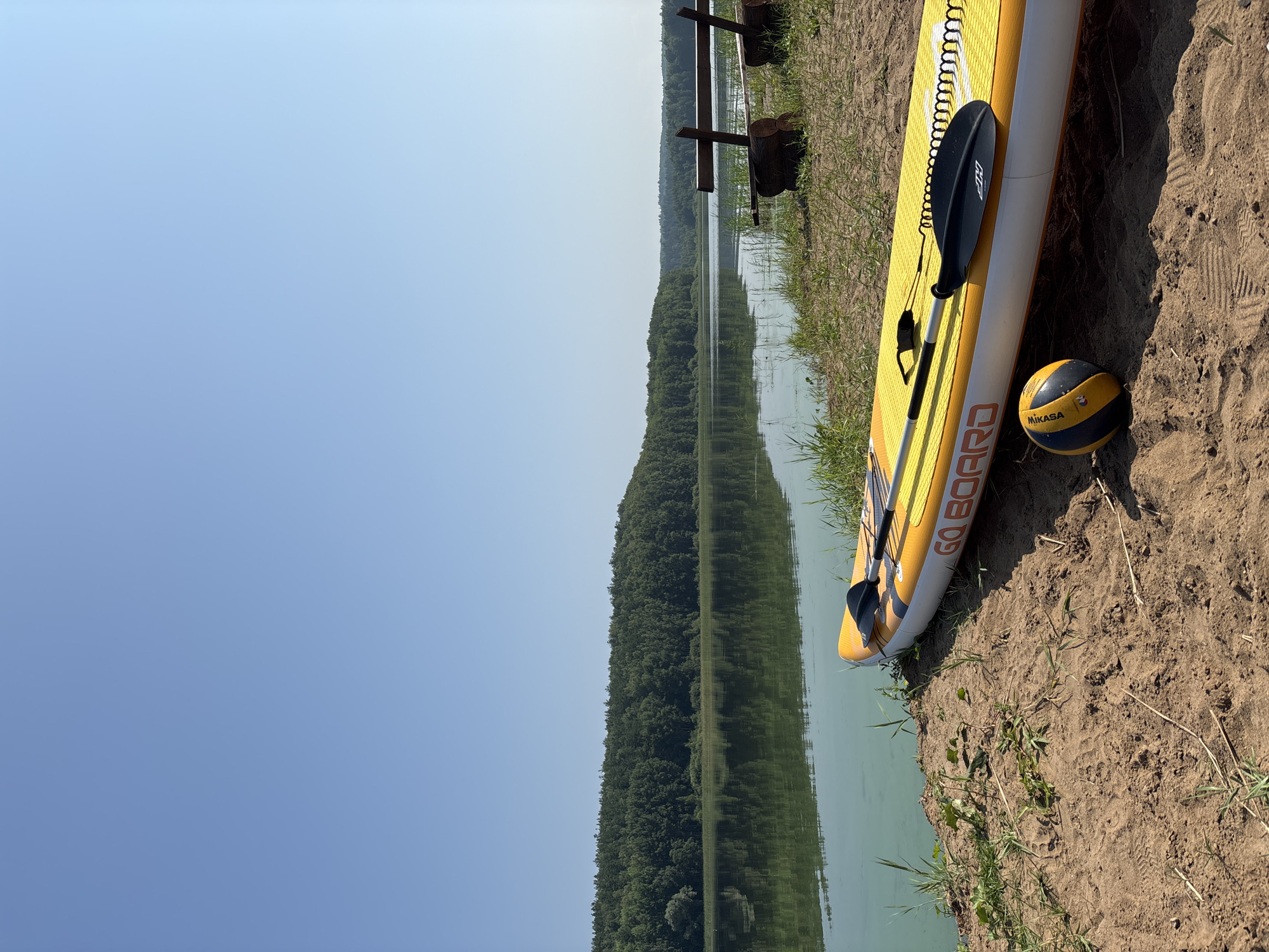 On a sunny day, a yellow and white stand-up paddleboard rests on a sandy shore next to a calm body of water. The paddleboard is equipped with a black paddle. A yellow and black volleyball sits on the sand in front of the paddleboard. In the background, a dense forest lines the opposite shore of the water, and the clear blue sky stretches above. Two rustic wooden benches are visible on the grassy area behind the sand. This scene is set in Chaykovsky, Russia, on the Kama River reservoir, also known as the Chaykovsky Pond.
