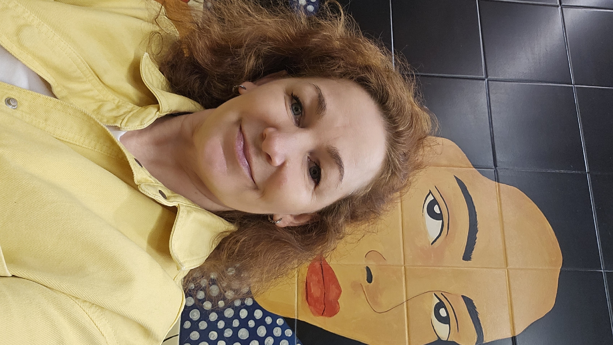 A woman with red curly hair, wearing a yellow shirt, is lying on a tiled surface. The surface features a cartoon-like face composed of tan tiles, black-tiled surroundings, and blue tiles patterned with white dots. The woman's head is positioned above the cartoon face's lips. Given that this image depicts a specific tile design in Sochi, Russia, the scene likely takes place in a public space or building featuring this unique decoration.