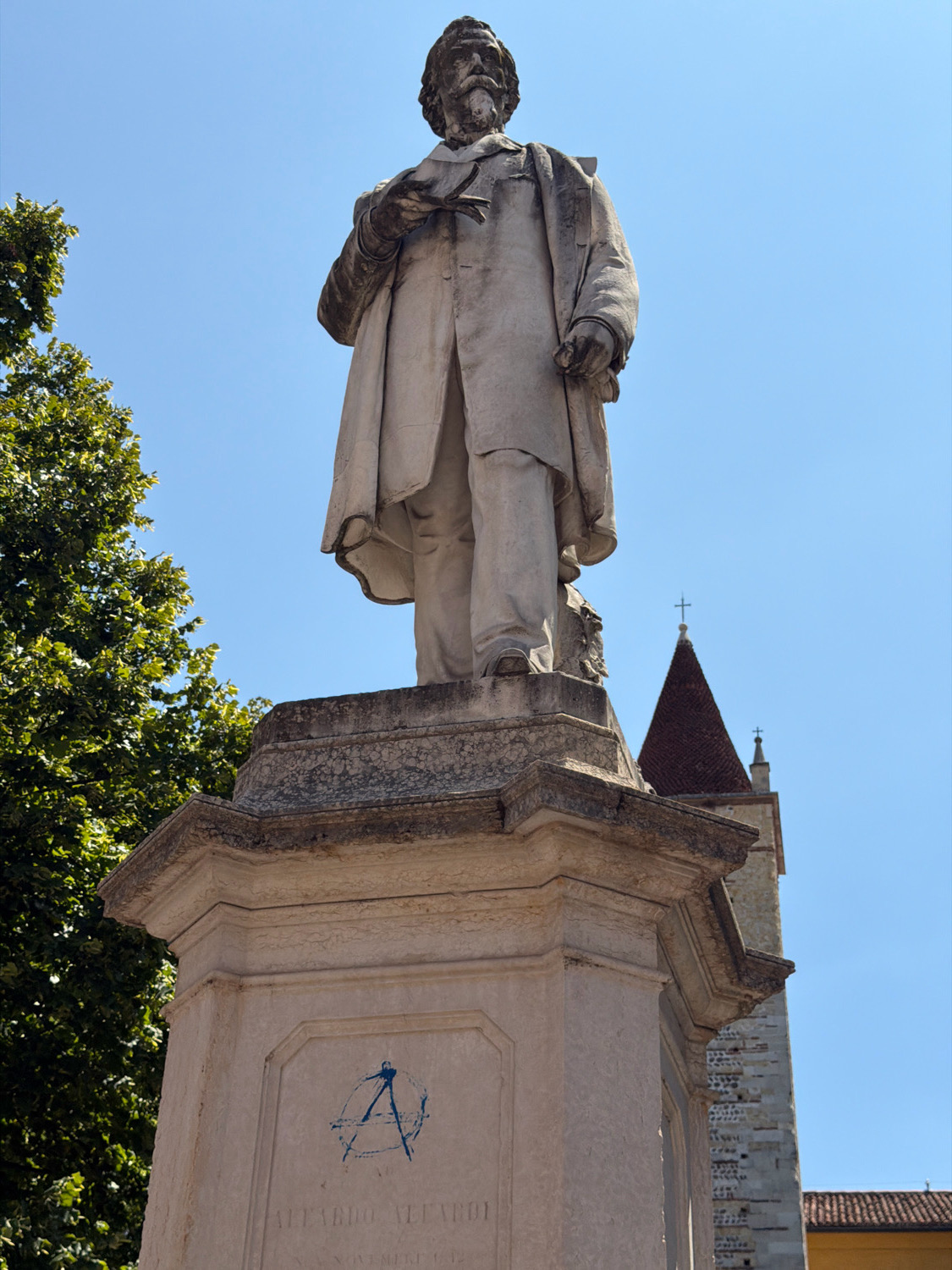 A low-angle, full-shot photograph captures a weathered stone statue of a man standing on a multi-tiered stone pedestal. The statue depicts the man from the waist up, wearing a long, draped coat and trousers. His beard is thick, and his hair is pulled back. He holds an object in his left hand and gestures with his right. The pedestal has a square base and features an inscribed name and date at the bottom. A blue anarchist symbol is spray-painted onto the front of the pedestal.
To the left of the statue, a large, leafy green tree dominates the frame. In the background, a tall stone tower with a reddish-brown conical roof and a cross on top rises into a clear blue sky. The architectural style of the tower suggests a historical European setting.
This statue is of the Italian poet and patriot Vittorio Alfieri and is located in Piazza Vittorio Veneto, Verona, Italy. The inscription on the pedestal reads "VITTORIO ALFIERI" and "XX NOVEMBRE MDCCCVII", marking his birth date. The anarchist symbol spray-painted on the monument is a recent addition, likely graffiti.