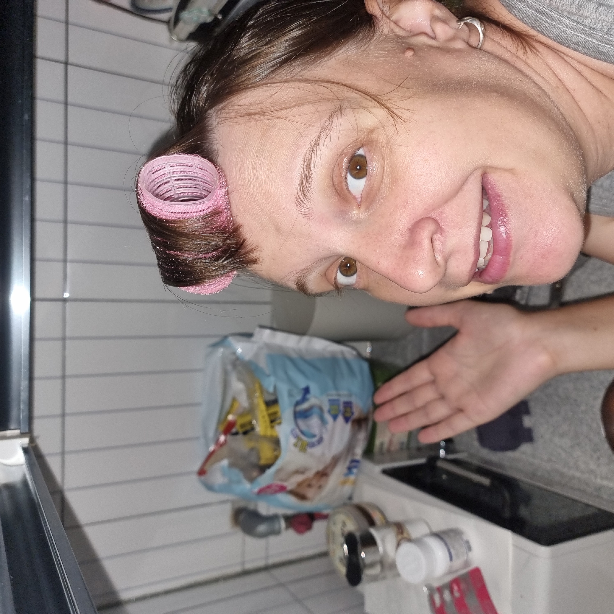A woman with brown hair styled with pink rollers is shown from the chest up in a close-up shot. She is smiling and looking directly at the camera, with her left hand raised and open, palm facing outward. She is wearing a grey shirt. Behind her, a white tiled wall is visible, along with various objects on a counter, including a blue bag filled with yellow items, jars, and a white bottle with a blue label. A faucet is also visible in the background. The overall setting appears to be a bathroom or kitchen area.
