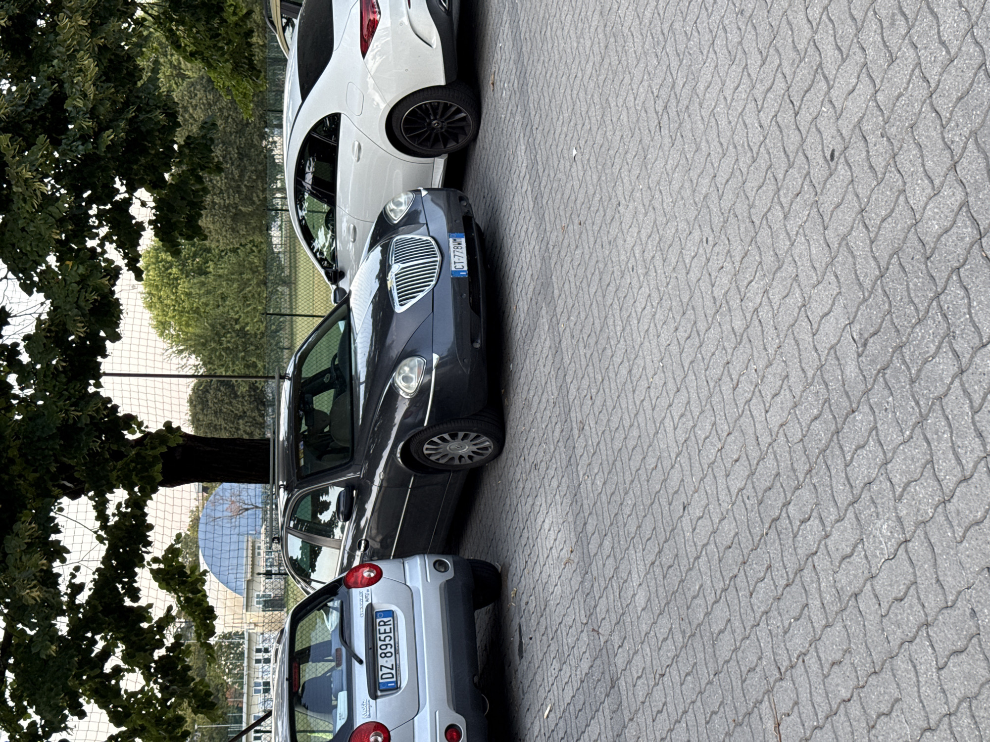 Three cars are parked on a cobblestone surface, with a large tree providing shade overhead. The cars are a silver Chevrolet Spark, a dark gray Lancia Thesis, and a white Mercedes-Benz CLA. The Chevrolet is on the left, the Lancia in the middle, and the Mercedes-Benz on the right. The Chevrolet has a license plate that reads "DZ 895ER". The Lancia has a license plate that reads "CT-778WH". In the background, there is a fence, some trees, and a building with a blue dome. The scene appears to be a parking lot in Parma, Italy, possibly near a sports facility or park, given the presence of the fence and net.