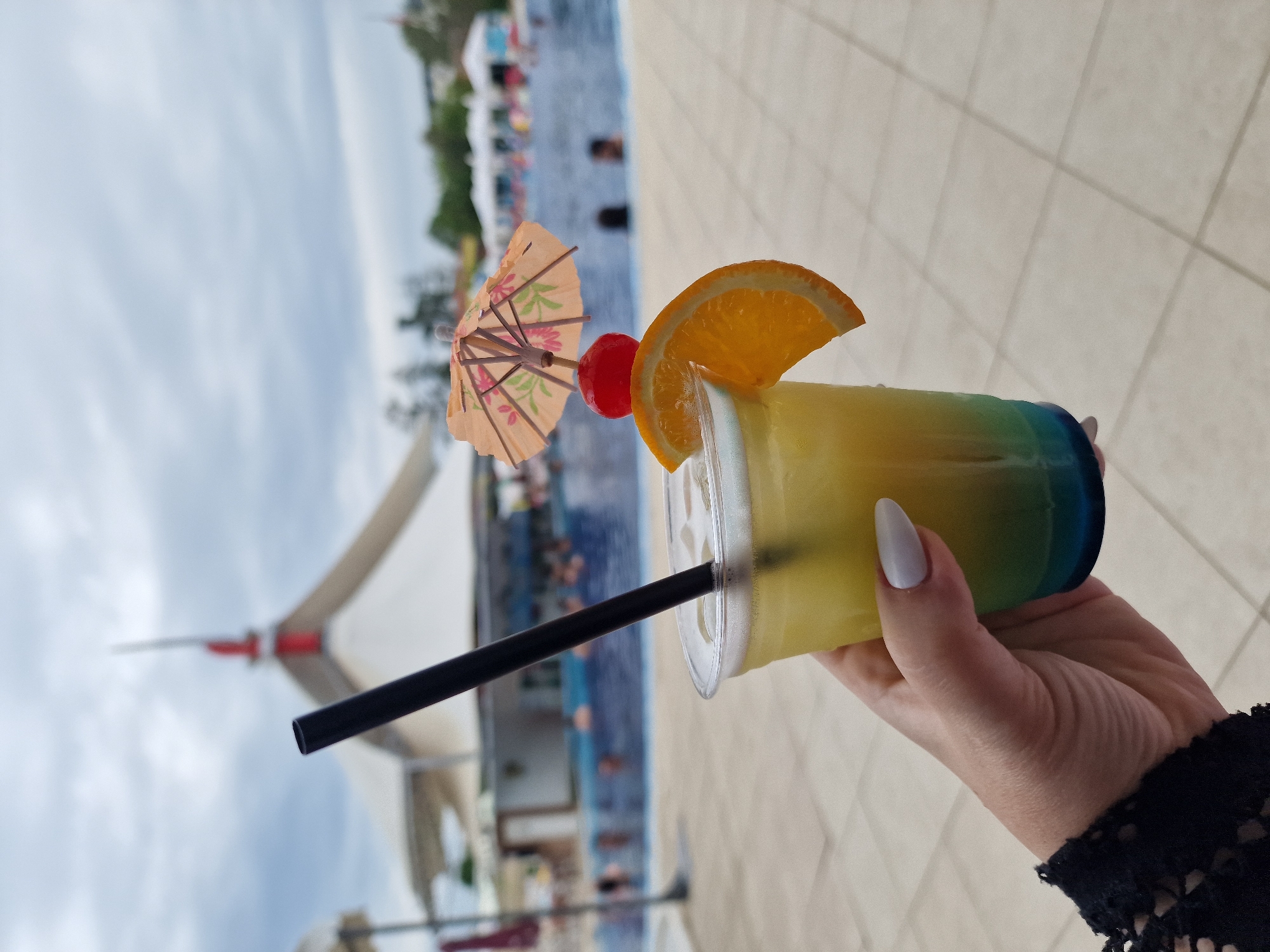 A hand with long, light-colored fingernails holds a clear plastic cup containing a layered, colorful drink. The drink is yellow at the top, fading to green, and then blue at the bottom. A black straw extends from the cup. The drink is garnished with a slice of orange and a red cherry, topped with a small, patterned paper umbrella. The background suggests an outdoor setting near a swimming pool, possibly a resort or recreational area. In the blurred background, there is a pool with people swimming, and a building with a white, tent-like roof. The ground appears to be paved with light-colored tiles. The sky is overcast. This scene is located at the Aqua Paradise swimming complex in Kazanlak, Bulgaria.