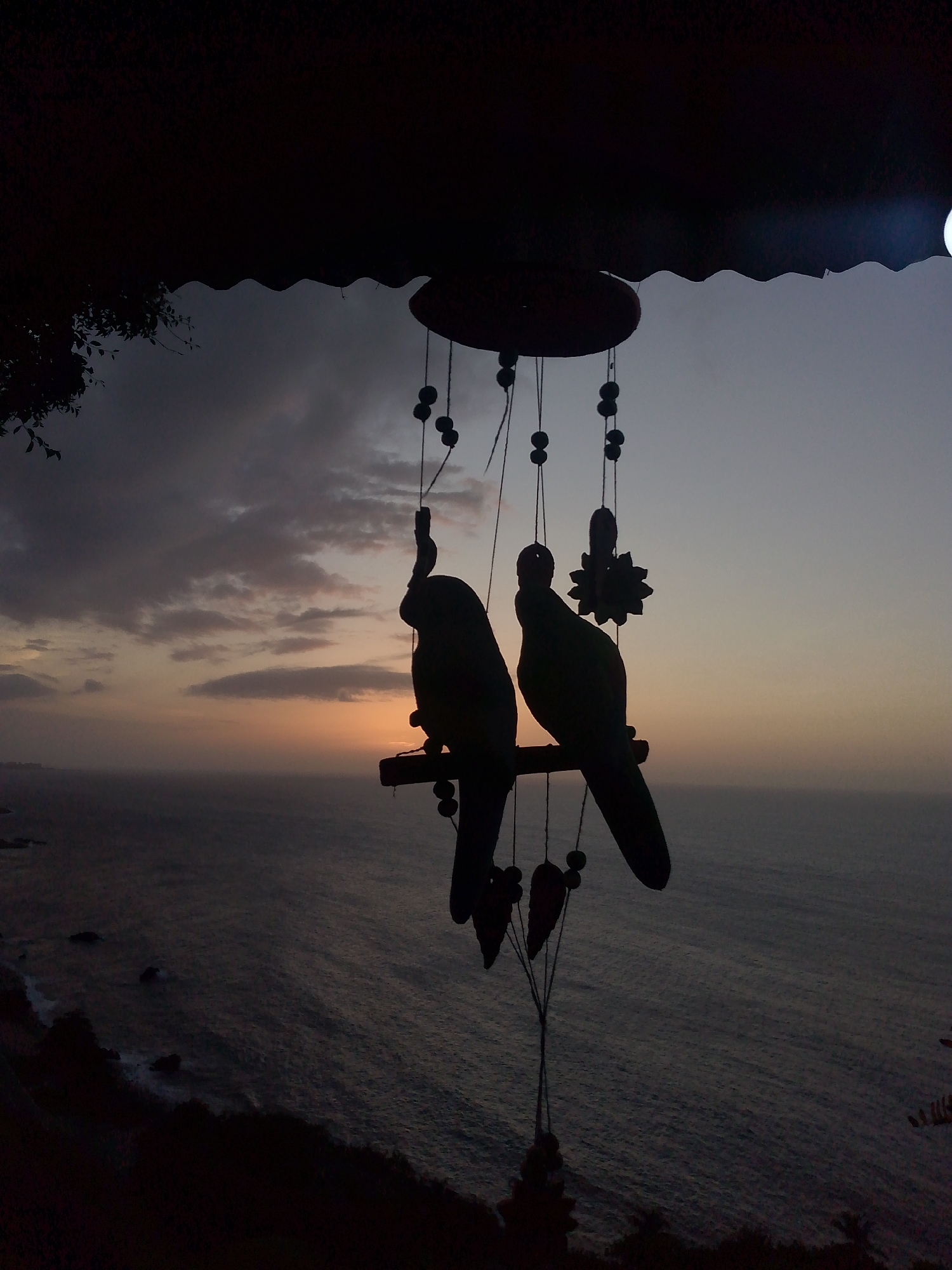 The image depicts a silhouette of two ornamental parrots hanging from a wind chime against a twilight sky over the ocean. The parrots are dark, with detailed shapes that suggest feathers and beaks. They are perched on a horizontal wooden dowel, which is part of the wind chime's structure. Several small, dark beads and a decorative floral element are also visible, hanging from threads connected to the top of the wind chime.
The background shows a calm ocean with gentle waves lapping at a distant shoreline, which is visible as a dark, undulating line against the water. The sky is a gradient of muted colors, transitioning from a pale orange and pink near the horizon to a dusky blue and gray above. Clouds are scattered across the sky, adding texture to the twilight scene.
In the foreground, a dark, silhouetted structure, possibly a roof or awning, extends from the top edge of the image, casting a shadow over the scene. To the left of the frame, a silhouette of foliage from a tree or bush is visible, further framing the view. The overall impression is one of tranquility and the serene beauty of dusk over a coastal landscape. The specific location in El Tigrillo, Venezuela, is not visually discernible from the image itself; it is provided as context.