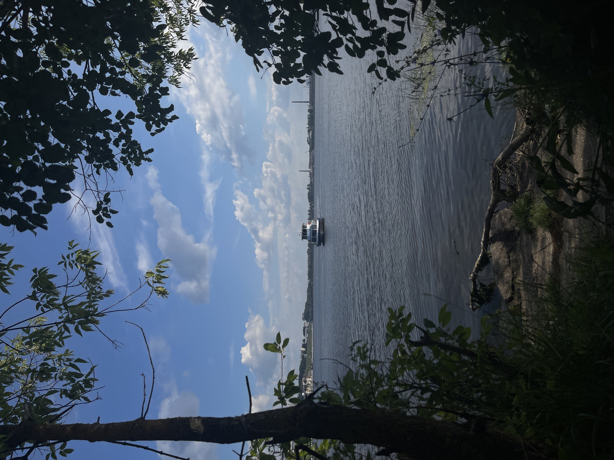 The image depicts a view of the Izhevsk Reservoir in Izhevsk, Russia, framed by trees and foliage in the foreground. A white boat is visible in the middle distance, moving across the calm water. The sky is blue with scattered white clouds. In the background, along the far shore, there are buildings and industrial structures, including chimneys. The lighting suggests it is daytime, possibly late morning or early afternoon.
