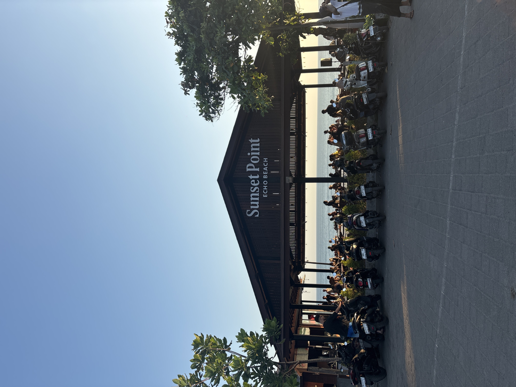 An eye-level, medium shot captures a view of the "Sunset Point Echo Beach" restaurant in North Kuta, Indonesia, as dusk approaches. The establishment, a dark-wood structure with a sloping roof, is set against a clear blue sky. The words "Sunset Point" are displayed in large white lettering above "ECHO BEACH." A row of motorcycles is parked in the foreground. The open-air restaurant is populated with numerous patrons seated at tables, many of whom appear to be enjoying the ocean view. The water stretches to the horizon, with a few figures visible along the shoreline. Green trees with dense foliage flank the left and right sides of the scene. The ground is paved with interlocking bricks.