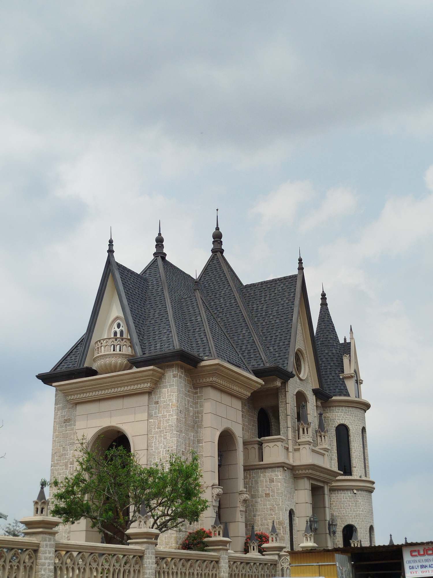 This media file captures a grand and ornate building, designed in a style reminiscent of a European castle or Gothic Revival mansion, set against an overcast sky. The primary subject is the multi-storied structure, characterized by its light beige and grey stone block facade and multiple steeply pitched dark grey shingled roofs. The roofs are adorned with numerous sharp spires and decorative finials.
Notable architectural details include an intricate balcony on the left side, featuring a delicate round window with a tracery pattern. The building also showcases several arched windows and doorways, decorative cornices, and detailed moldings that emphasize its elaborate design. On the right, a cylindrical tower with tall, narrow, dark-paned windows adds to the castle-like appearance.
In the foreground, a decorative light-colored stone or concrete fence with intricate, gothic-inspired patterns partially obscures the lower section of the building. A vibrant green leafy tree is visible in front of the building on the left, and a small patch of red flowering plants adds a touch of color near the fence.
The scene is set during the daytime, likely late morning or early afternoon, under a cloudy sky with soft, scattered clouds, indicating an overcast day. There is no human activity or event taking place; the image focuses solely on the architecture and its immediate surroundings. The presence of such a European-styled building in Da Lat, Vietnam, serves as a significant location cue.
In the bottom right corner, a partial view of a simple structure with a corrugated metal roof reveals a red sign with Vietnamese text. The visible text includes: "CỬA" (likely part of 'Cửa hàng' - shop), "CHUYÊN SỬA ĐIỆN CƠ" (Specializing in electric motor repair), "ĐẠI LÝ BÌNH ACQ" (Binh ACQ agent/dealer), and a phone number starting "ĐT: 0917...".