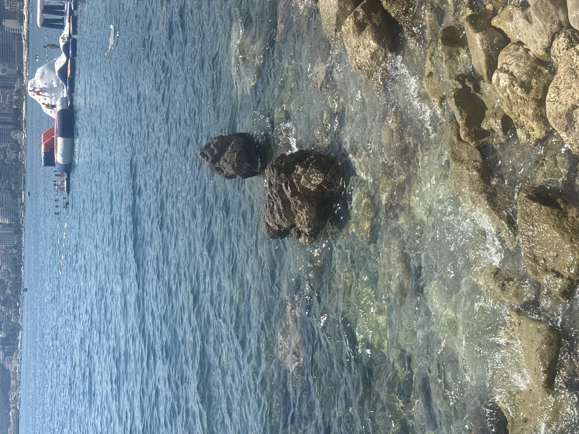 The image depicts a sunny day at a rocky beach in Budva, Montenegro. In the foreground, numerous natural rocks of varying sizes are partially submerged in clear, shallow water. Further out in the sea, an inflatable water park with slides and climbing structures is visible. Several individuals are interacting with the water park, including people climbing on a large white inflatable structure and others swimming in the water. A person is also swimming in the sea closer to the foreground. In the background, a cityscape with buildings and a hazy horizon suggests the urban environment of Budva. The water is calm with small waves lapping against the rocks.