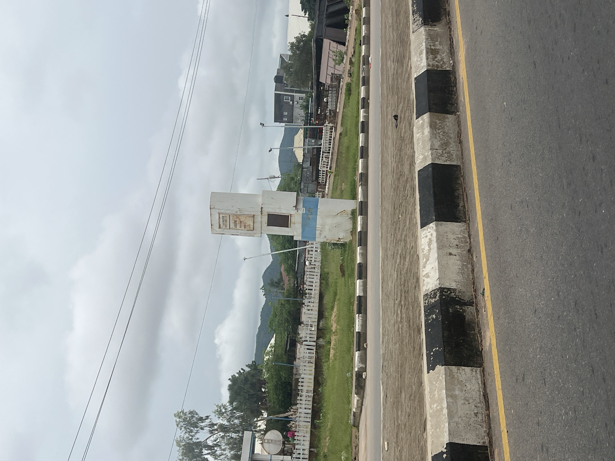 The image shows a cloudy day in Jalingo, Nigeria, near a road. In the foreground, a paved road with a yellow line is visible. A concrete curb with alternating black and white painted sections runs along the side of the road. Beyond the curb, there is a grassy area with a white picket fence. To the left, a white cylindrical structure with a blue stripe is partially visible, possibly a billboard or a signpost. Further back, there are trees, buildings, and hills in the distance. Some power lines are strung across the sky.