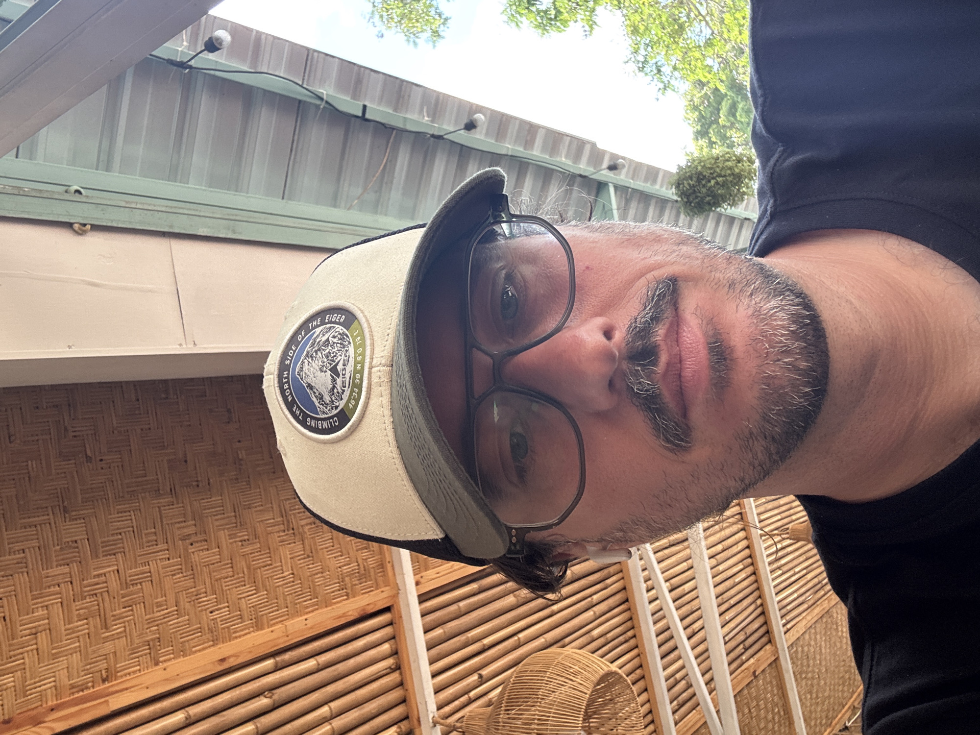 A photograph depicts a man wearing a baseball cap and glasses. The location is Kuta Selatan, Indonesia. The surrounding environment includes a woven bamboo wall and roof.