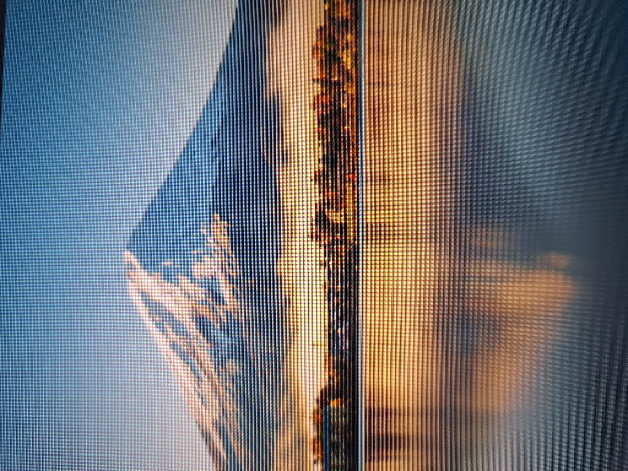 The image displays a serene landscape dominated by a majestic mountain, likely Mount Fuji, capped with snow and bathed in the warm glow of sunrise or sunset. The lower portion of the image features a calm body of water that perfectly mirrors the mountain and the sky, creating a sense of symmetry and tranquility. A distant treeline with autumnal foliage separates the mountain from the water. The sky above is a clear, soft blue, transitioning to warmer hues near the horizon where the sun's light illuminates the clouds. The scene evokes a sense of peace and natural beauty. This image does not depict any location in the city of Varna, Bulgaria.