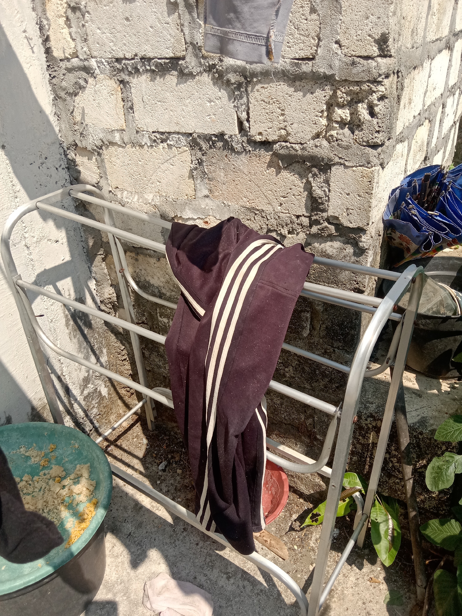 A dark-colored garment with three white stripes is hanging on a metal drying rack placed against a rough, textured brick wall. The drying rack is silver and appears to be made of lightweight metal tubing. To the left of the drying rack is a green plastic basin filled with a yellowish, granular substance. A blue umbrella with its handle pointing upwards is leaning against the wall to the right of the drying rack, partially obscuring a black bucket. Green leaves from a nearby plant are visible in the lower right corner of the image. The scene is lit by bright sunlight, casting shadows on the ground and wall. The setting appears to be outdoors, possibly in a residential area in Stiaji, Indonesia, indicated by the characteristic brick wall and the presence of household items.
